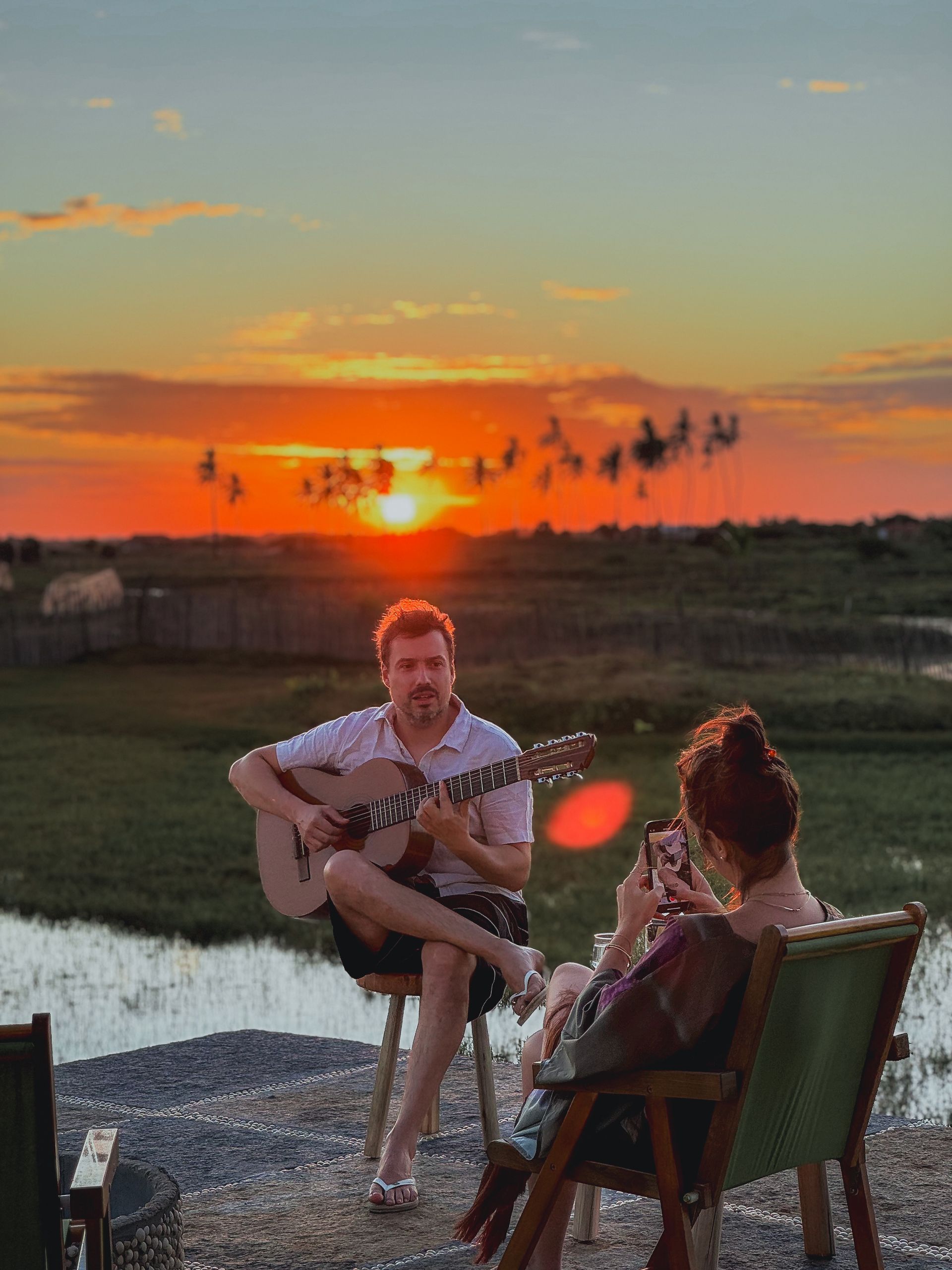 Uma pessoa toca violão para outra sentada em uma cadeira, tendo como pano de fundo um vibrante pôr do sol alaranjado sobre um campo e a água.