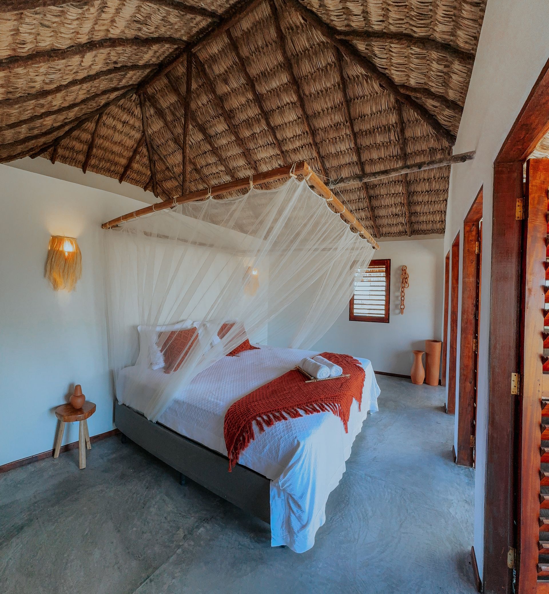 Quarto com teto de palha, cama com dossel, roupa de cama branca, manta laranja, persianas de madeira e banquinho.