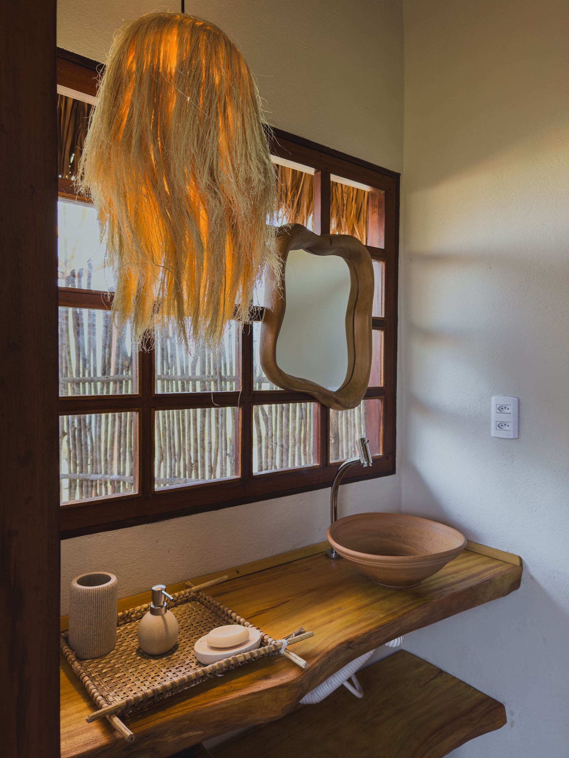 Banheiro com pia de madeira, espelho e luminária perto de uma janela com teto de palha.