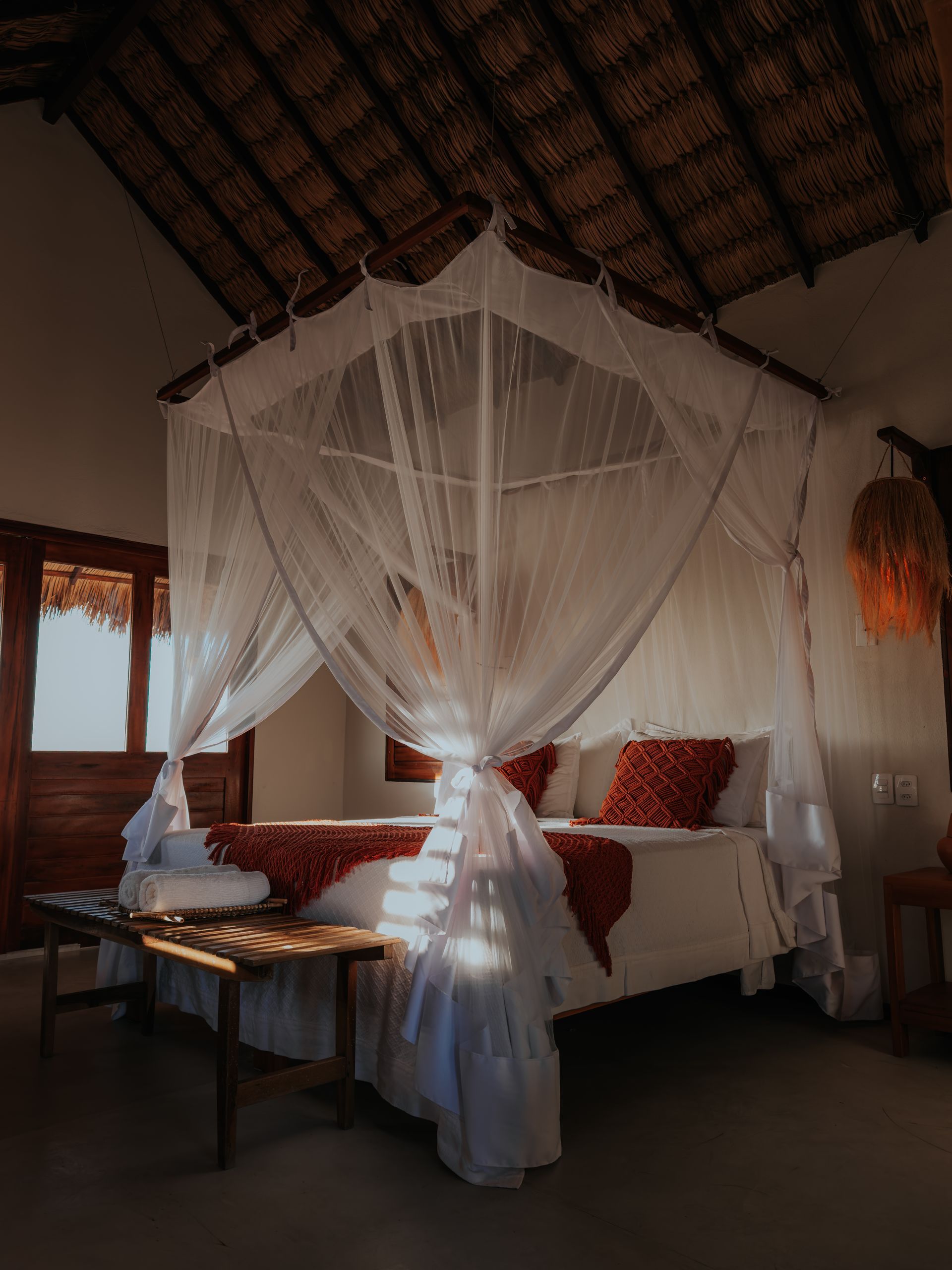 Cama com mosquiteiro em quarto ensolarado, teto de palha, detalhes em vermelho na roupa de cama, móveis de madeira.
