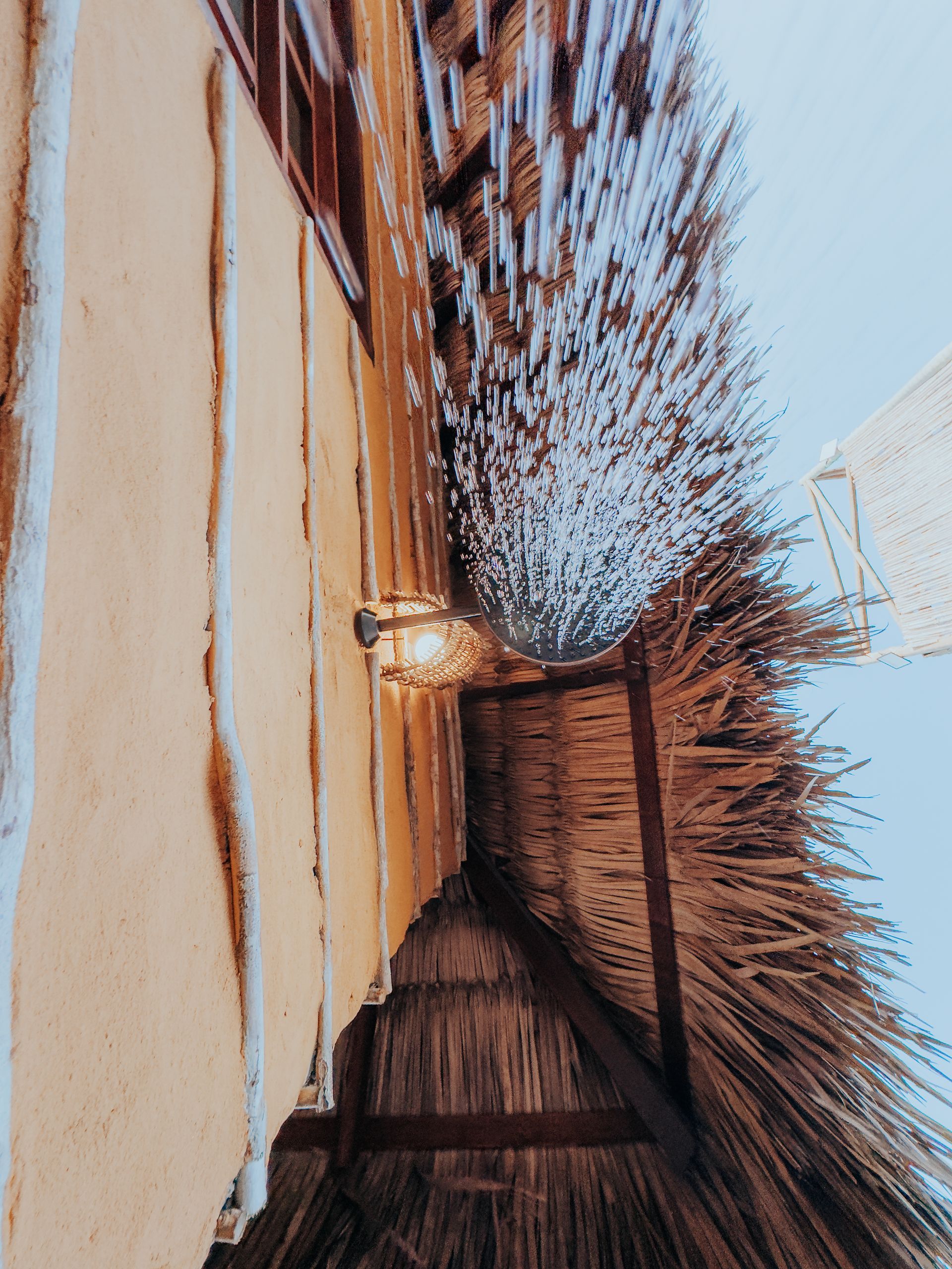 Vista de um telhado de palha com uma luminária em uma parede de estuque de cor clara contra um céu azul.