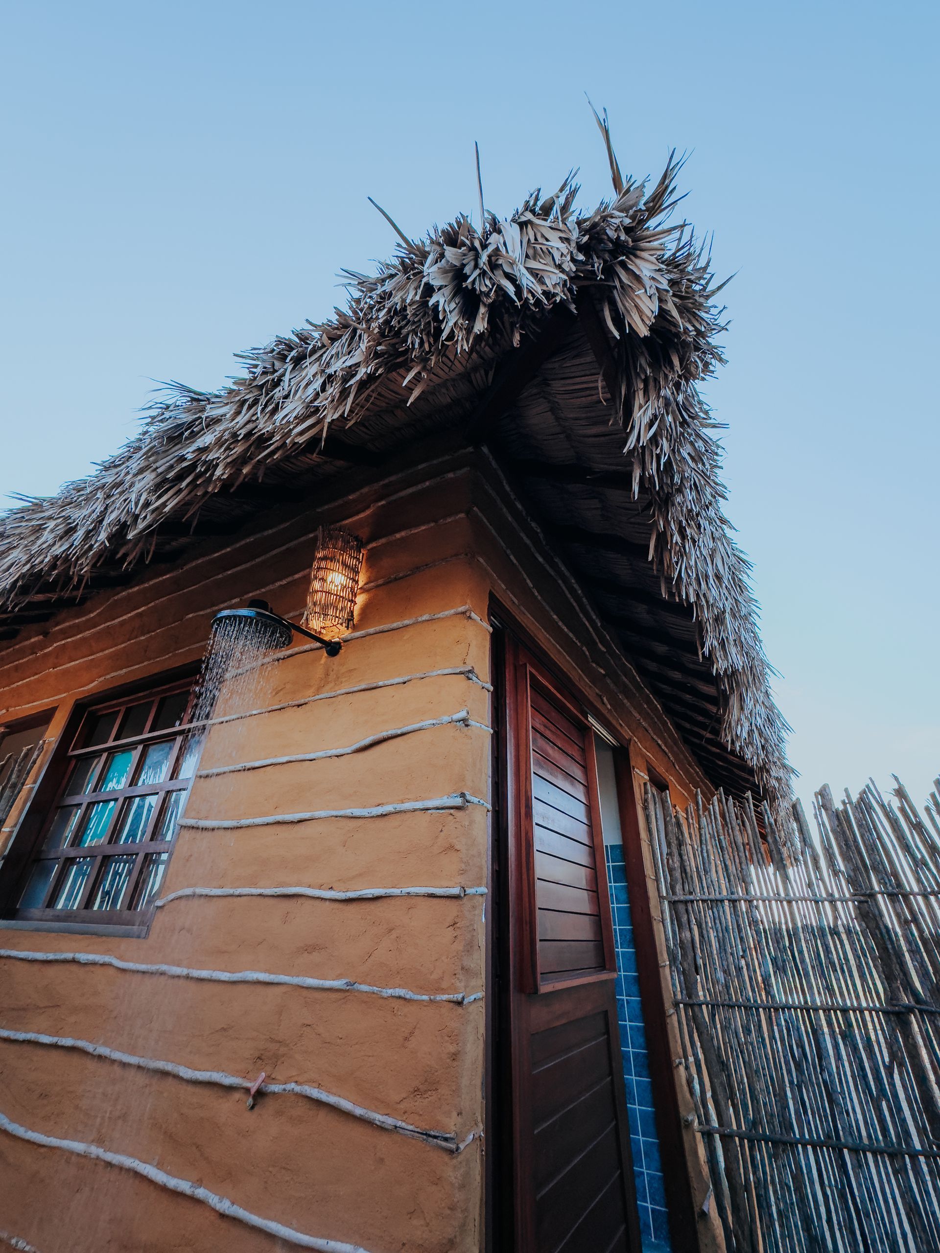 Cabana de madeira com telhado de palha, iluminada por um aplique de parede, contra um céu azul claro.
