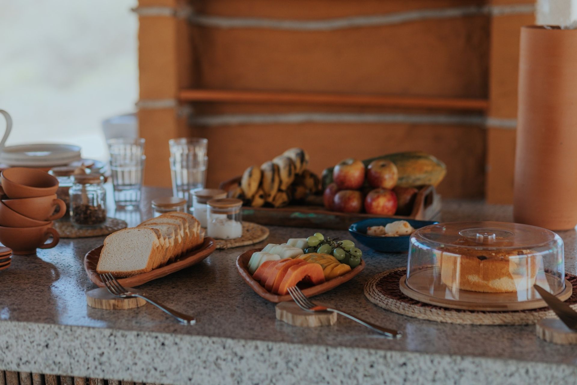 Buffet de café da manhã: pão, frutas, bolo, xícaras e copos dispostos em um balcão com fundo de tijolos de barro.