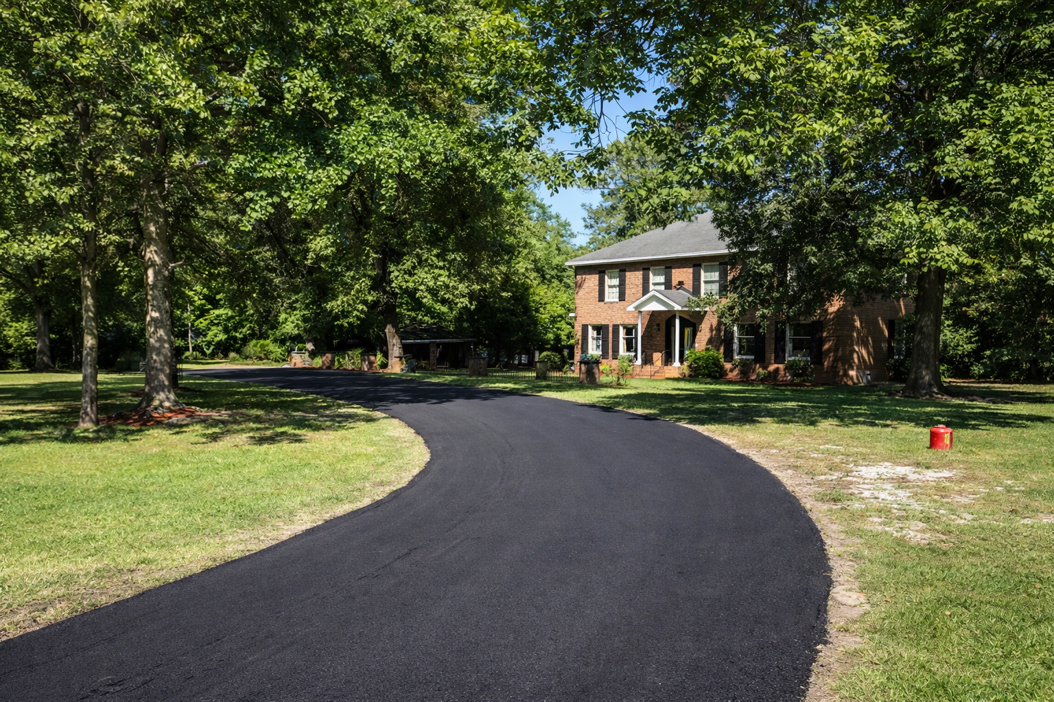 A freshly paved black asphalt driveway curves through a lush green yard towards a house sheltered by trees.