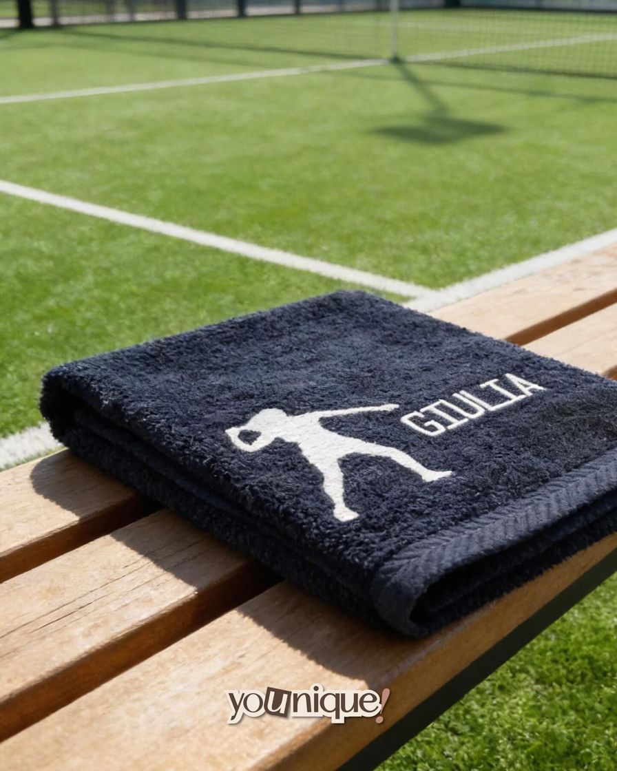 Asciugamano nero GILLIA con logo bianco su una panchina di legno accanto a un campo sportivo verde