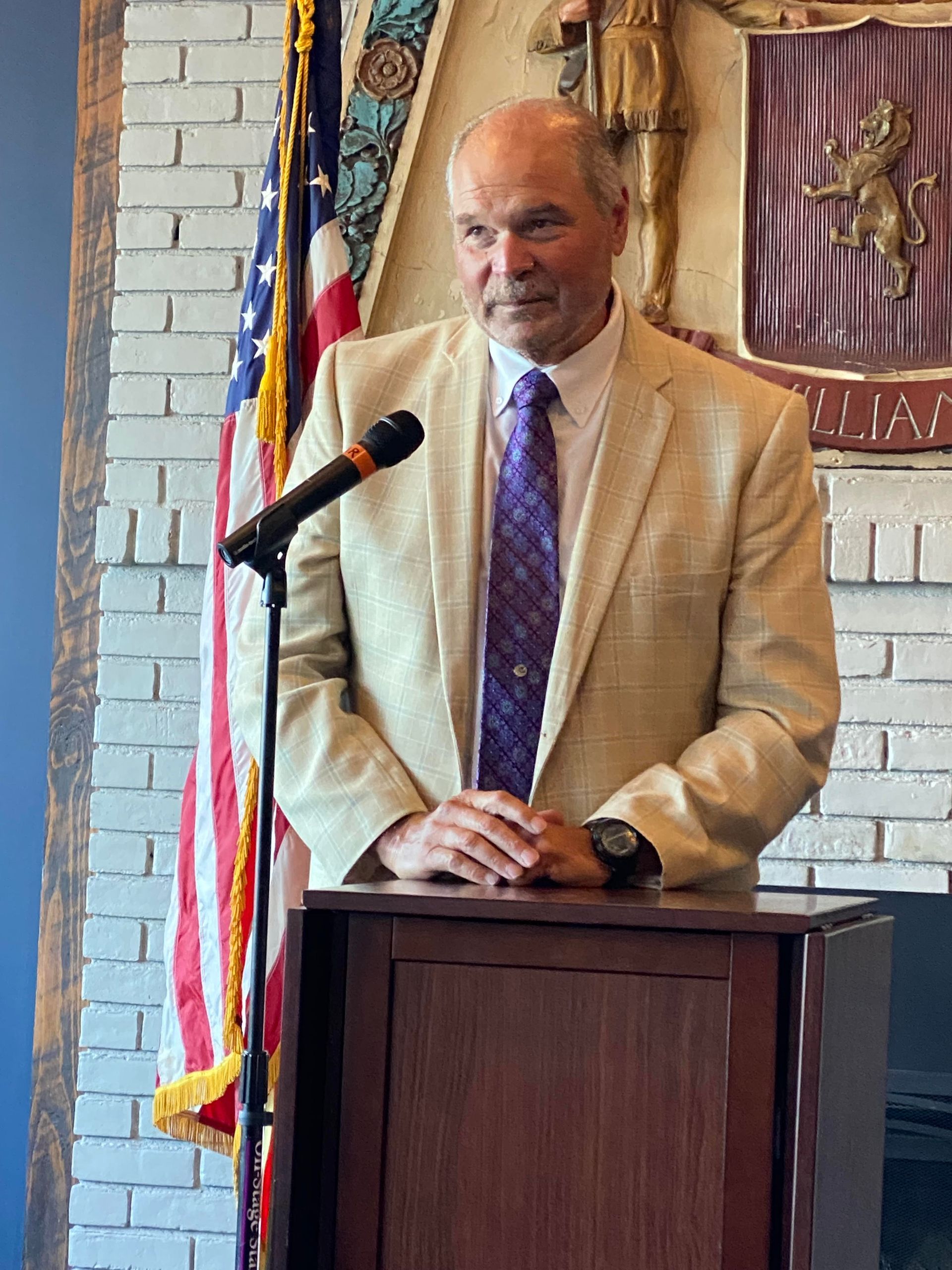 Hon. Judge Robert Muller (Ret.) in blazer speaks at a podium. American flag and crest in the background.