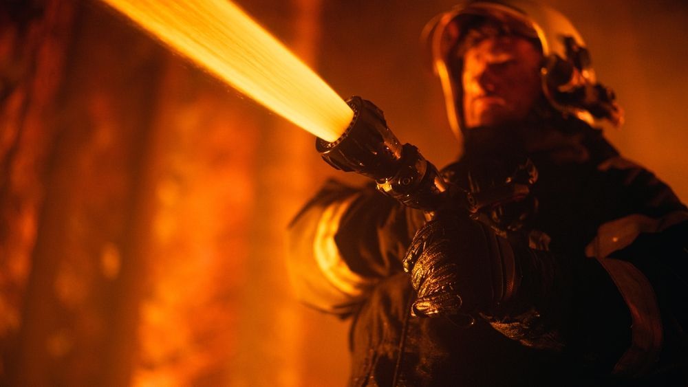 Firefighter in black gear aims a hose, blasting bright orange water or foam in a smoky scene