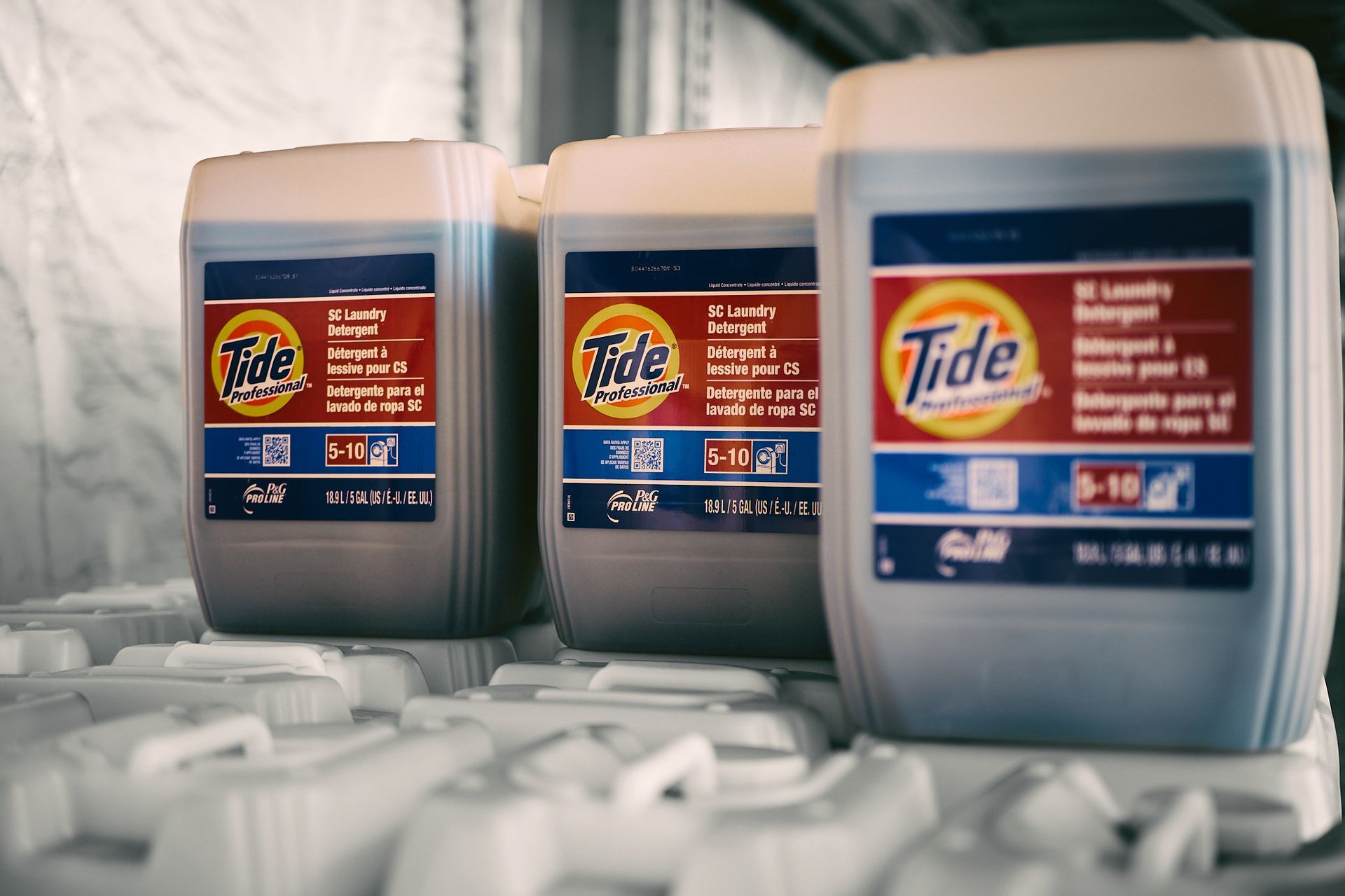 Three large Tide laundry detergent containers on a shelf. Red and blue labels, beige containers.