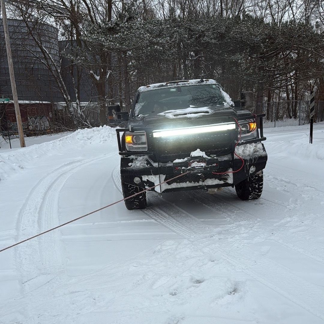 A dark pickup truck with a glowing grille light bar through a snow-covered outdoor area doing a winch out