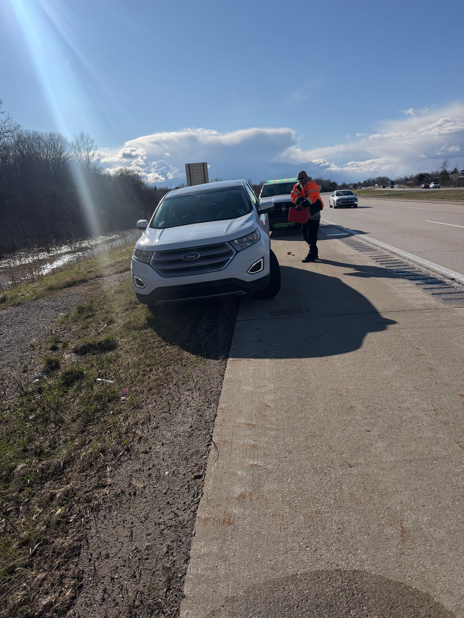 A white SUV parked on the shoulder of a highway, with an individual in an orange high-visibility vest standing nearby.
