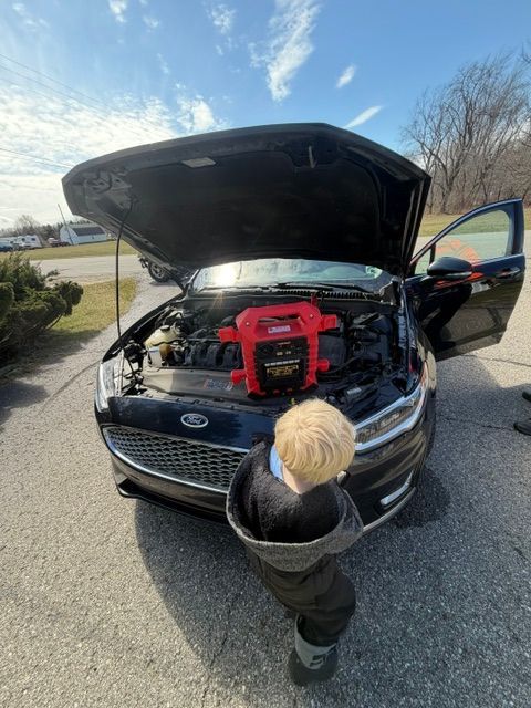 A child stands in front of a Ford vehicle with an open hood, on which a red portable car jump starter is placed.