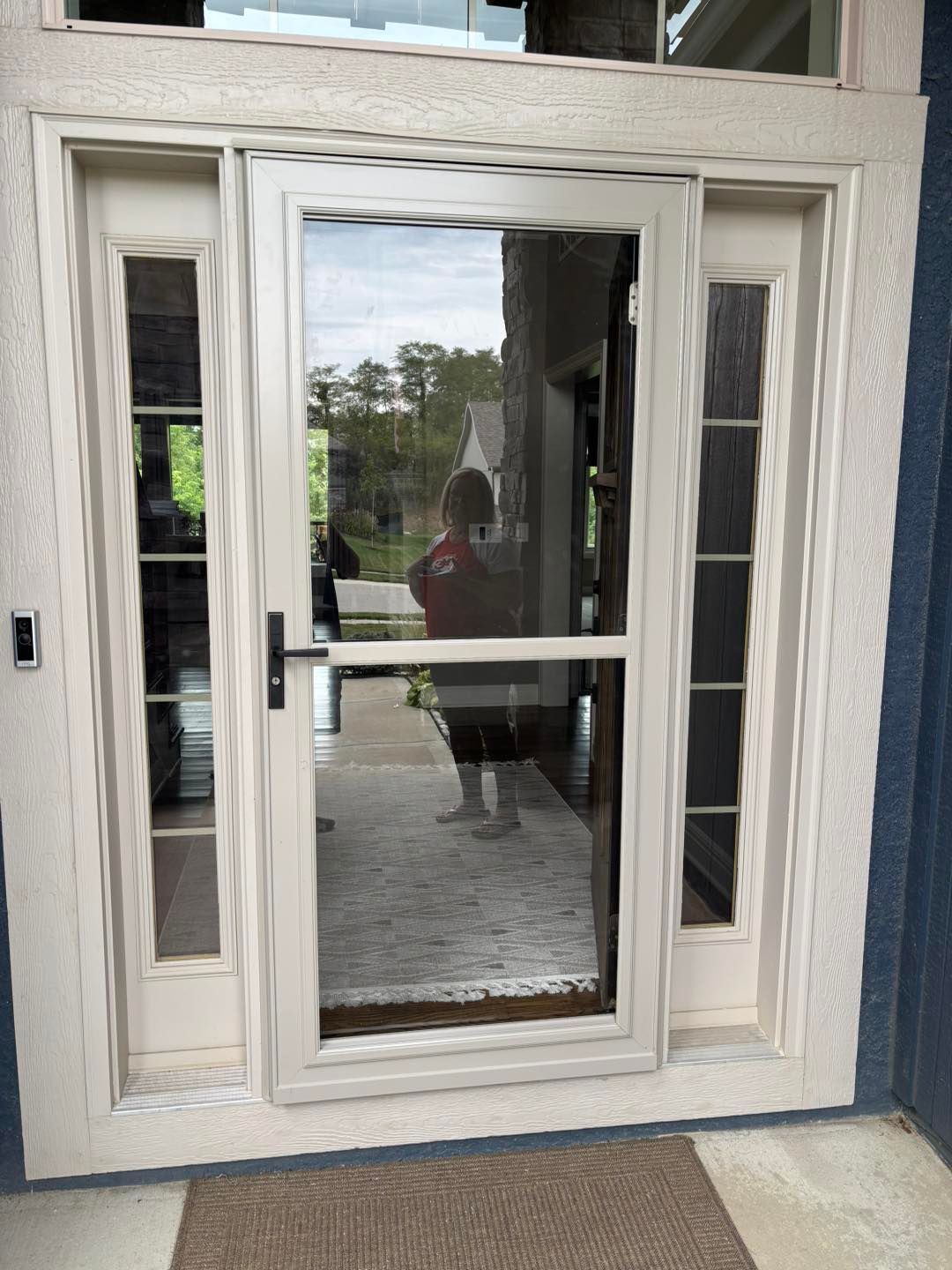 Screen door with a person reflected, flanked by glass panels, cream trim, and a doormat.