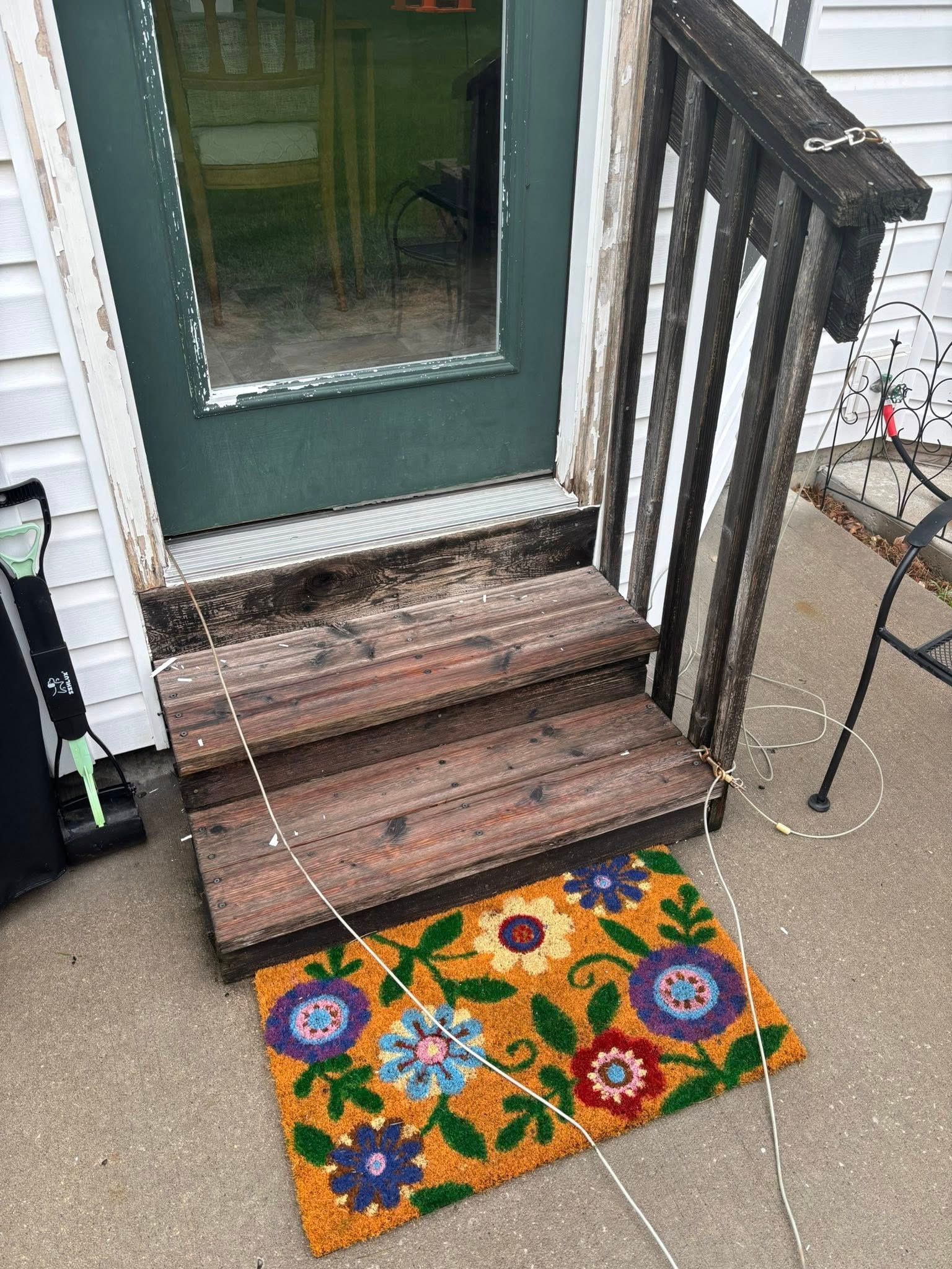 Wooden steps leading to a green door with a floral doormat on concrete.