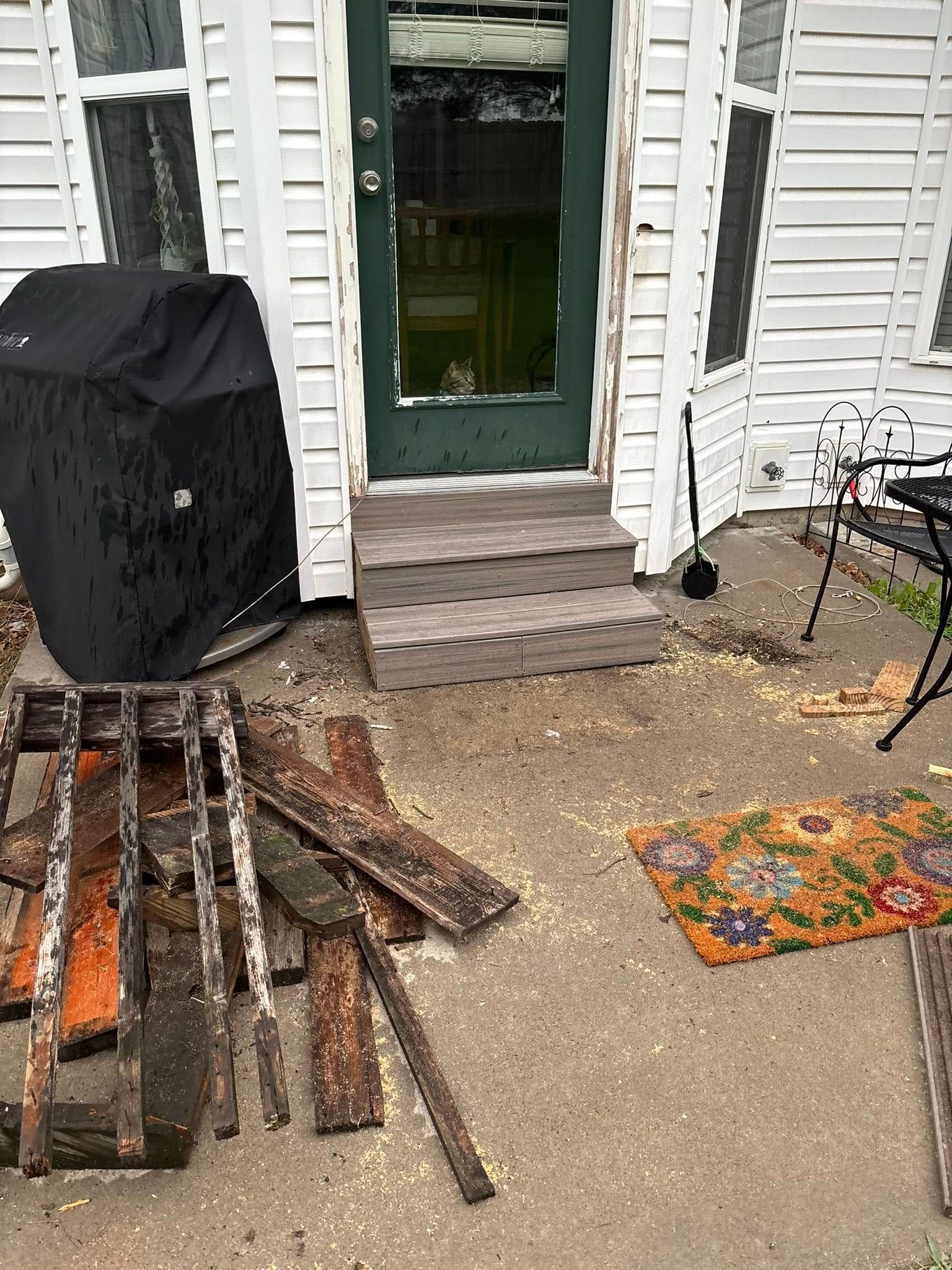 Debris and a doormat clutter the concrete entrance to a white house with a green door.