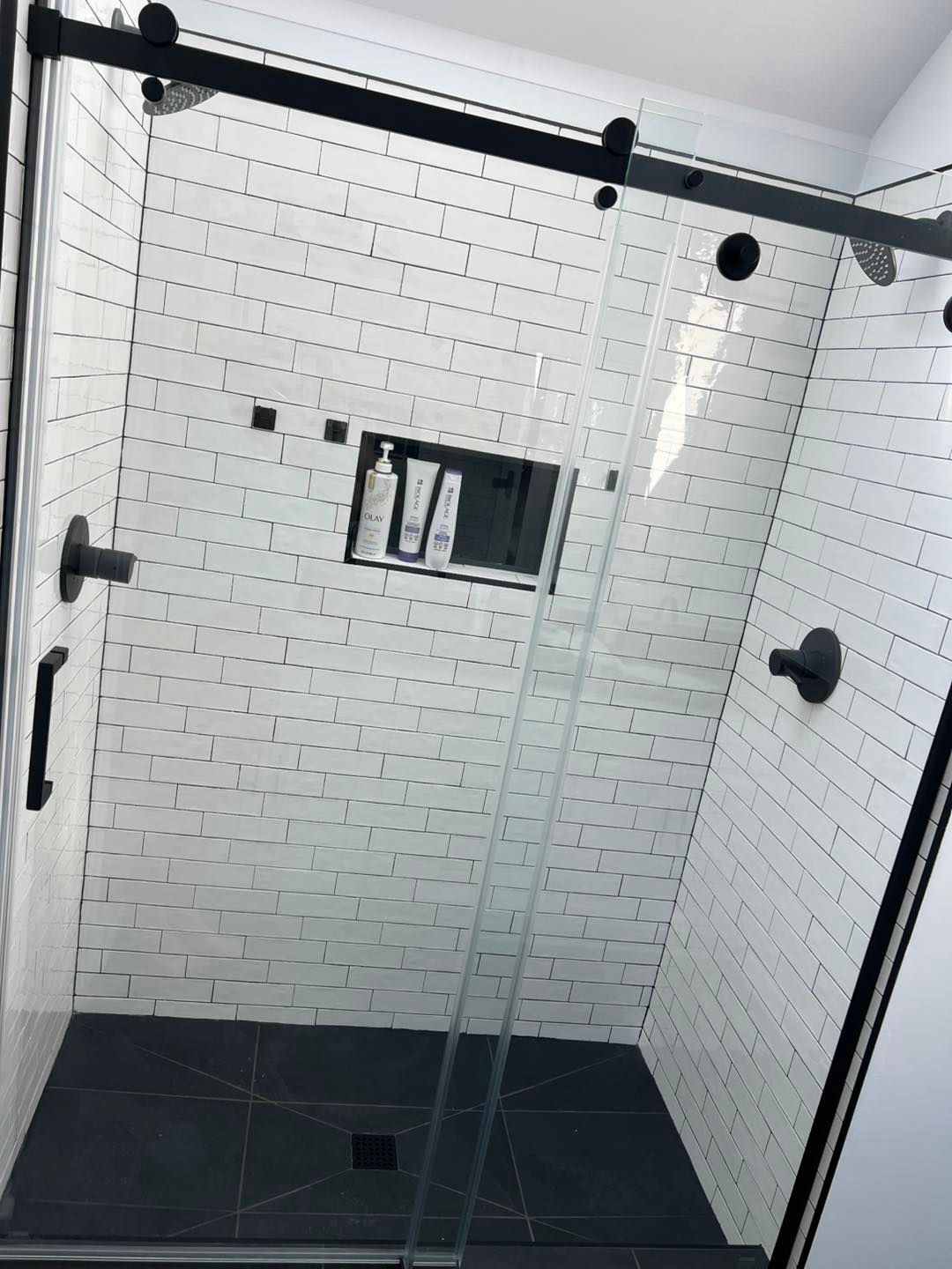 Black-framed glass shower enclosure with white brick-like tiled walls and a dark gray tiled floor.