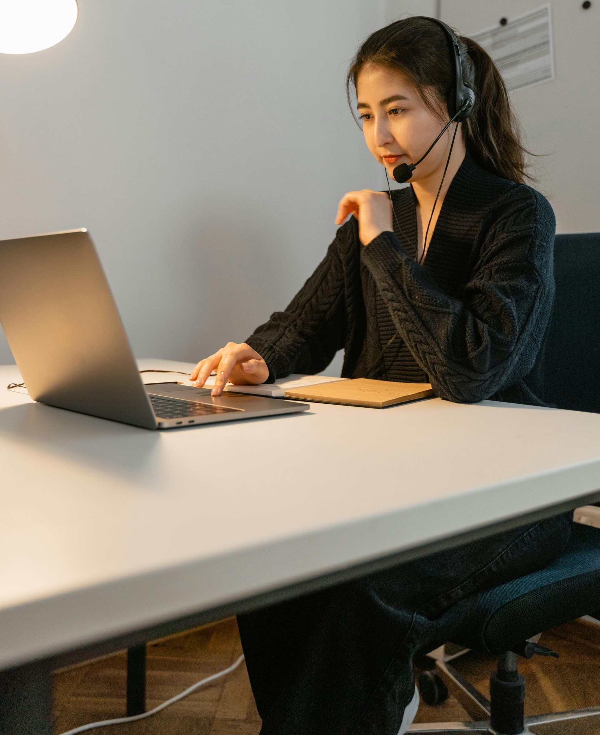 Woman wearing headset, working on laptop at desk. Office setting.