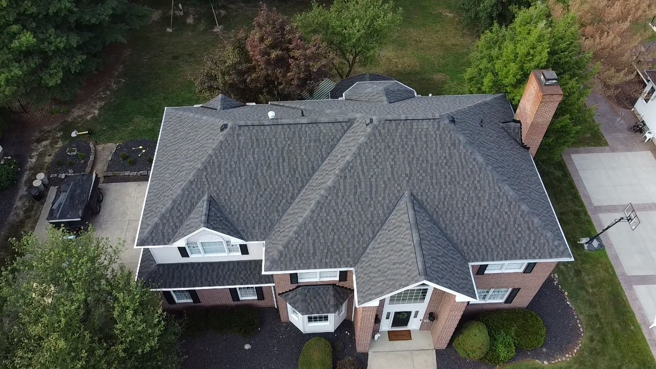 Aerial view of a gray-roofed brick house with a driveway and surrounding trees.