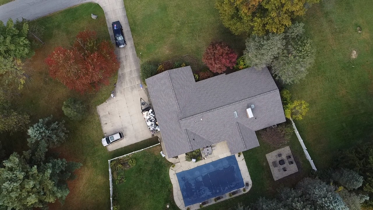 Aerial view of a house with a blue pool, driveway, lawn, and trees surrounding it