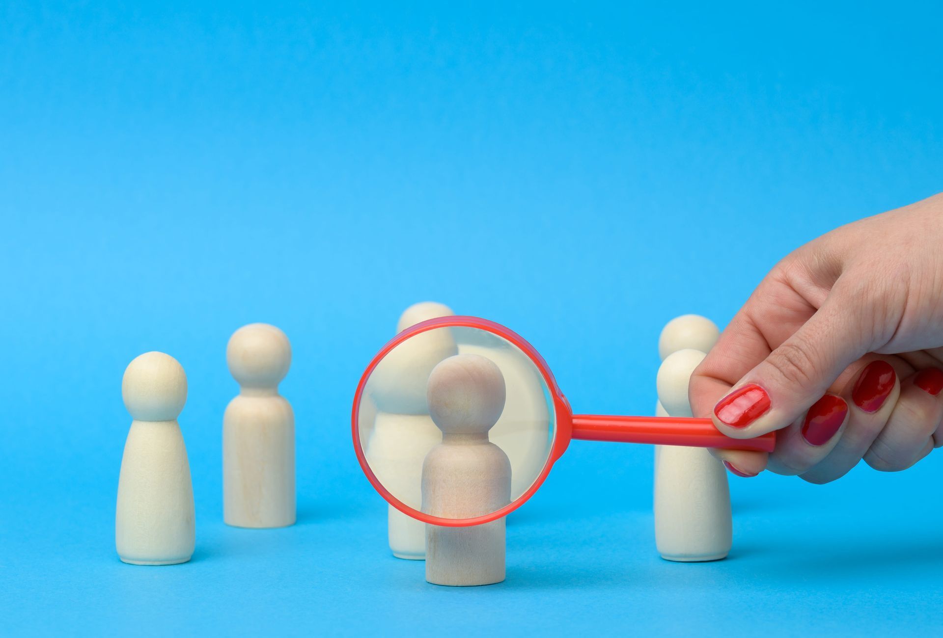 Hand holding a magnifying glass over a central wooden figure among several others on blue background