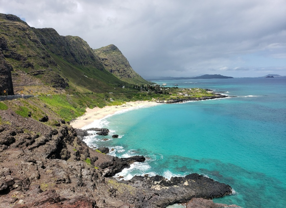 Turquoise bay with white sand beach, rocky green cliffs, and cloudy sky