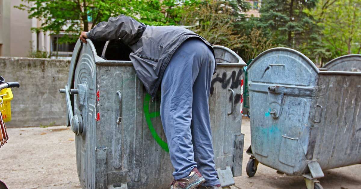 A man in blue jeans and a gray coat is leaning into a dumpster. His legs are hanging out of it.