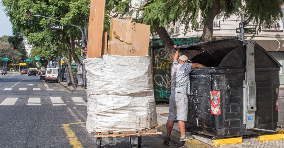 Man with a flat trolley filled with findings is searching a dumpster on the open street.