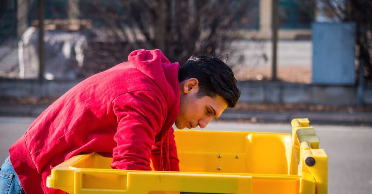 A young man who is wearing a red hoodie is leaning over and looking into a bright yellow dumpster.