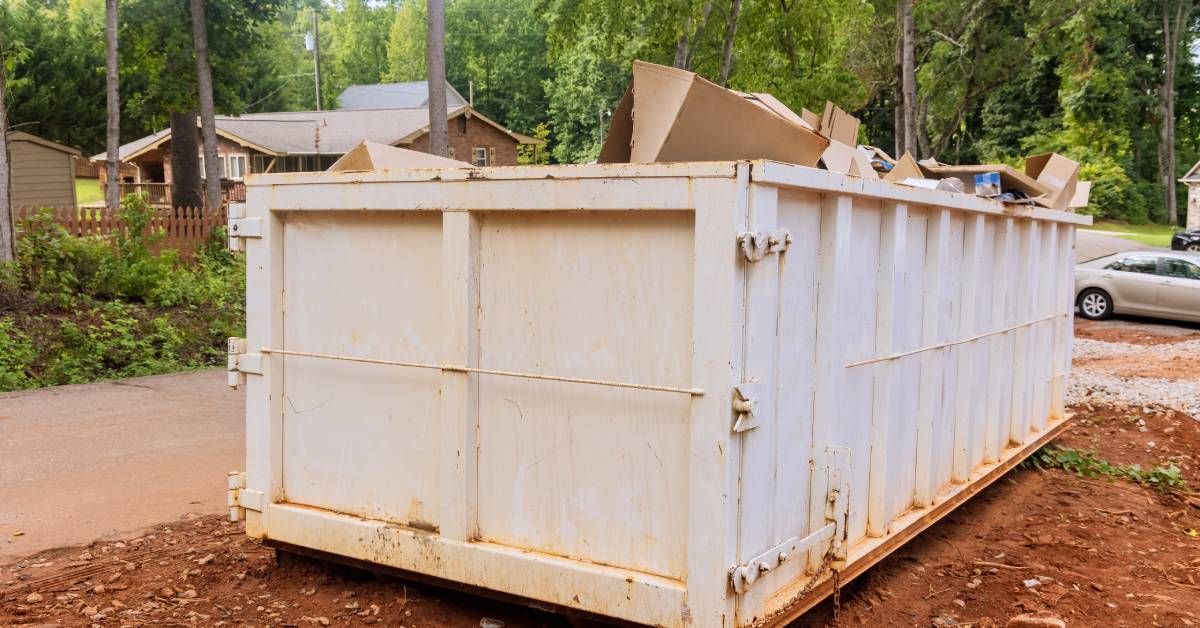 A large beige dumpster unit parked on a yard. The dumpster is overflowing with cardboard and other debris.