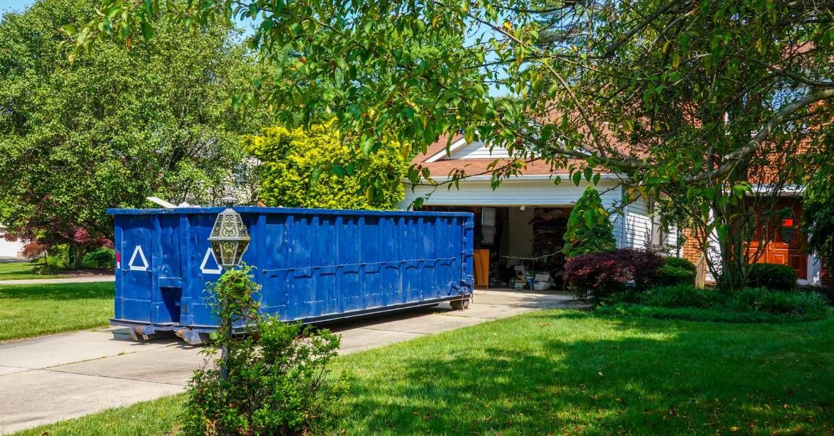 A large blue dumpster in the driveway of a residential house. The garage door is open, showing that it is full of stuff.