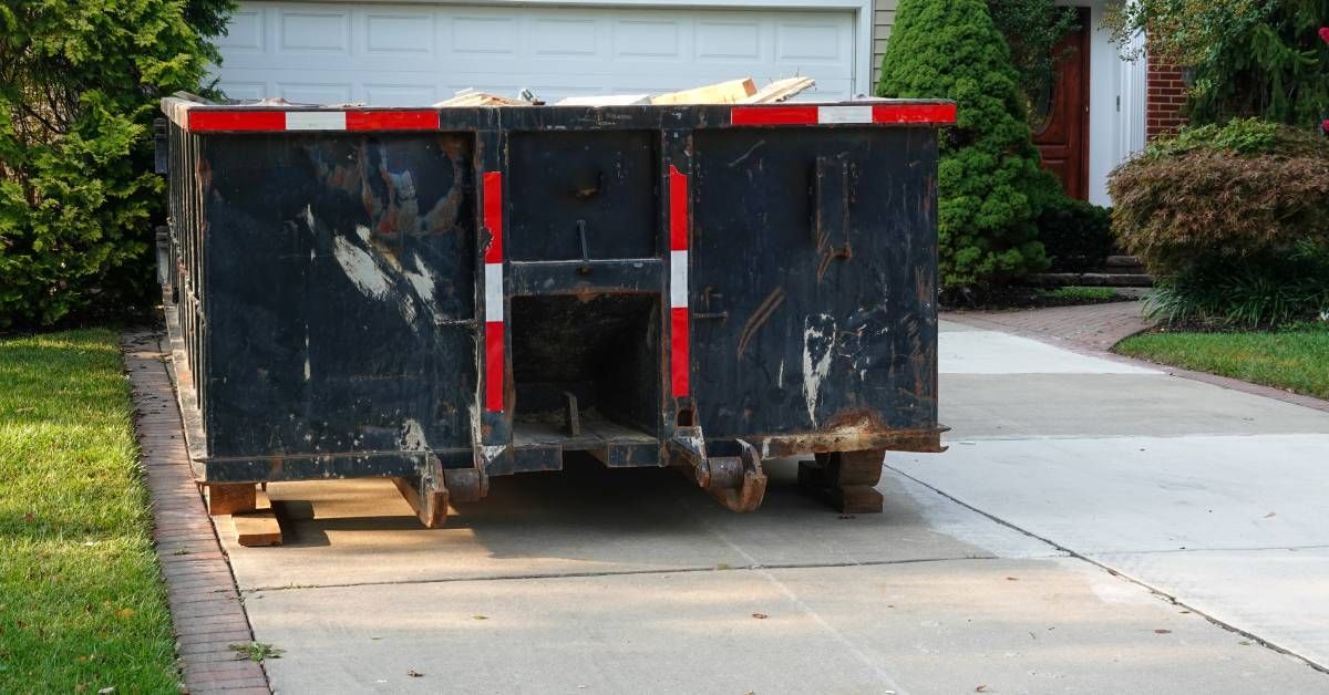 A long, black dumpster parked in the concrete driveway in front of a residential house.