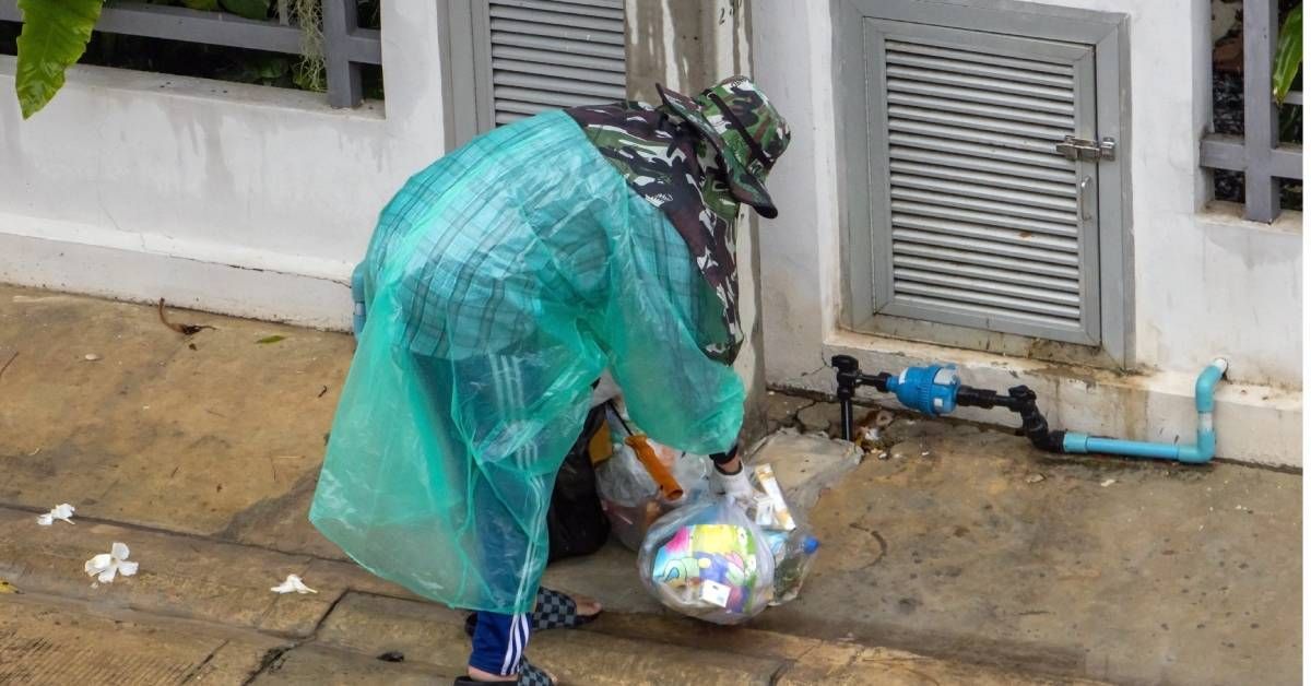 A person in camo clothes and poncho is out on the street. They're collecting garbage bags in the rainy weather.