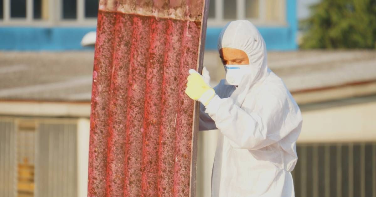 A man in full PPE carrying a large wavy sheet of roofing material with a red underside.