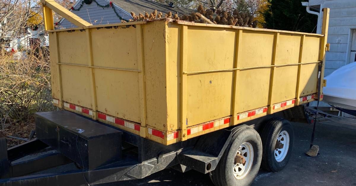 A yellow dumpster unit on a black trailer in a driveway. The dumpster is full of wood sticking out of the top.