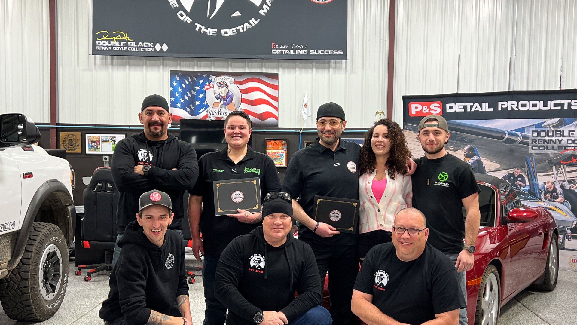Group of people in a shop, some holding certificates. Red car and white truck on sides.  American flag art.