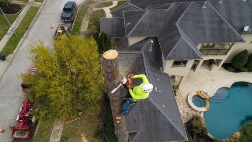 A person in a harness, using a chainsaw on a tree trunk atop a house roof near a pool.
