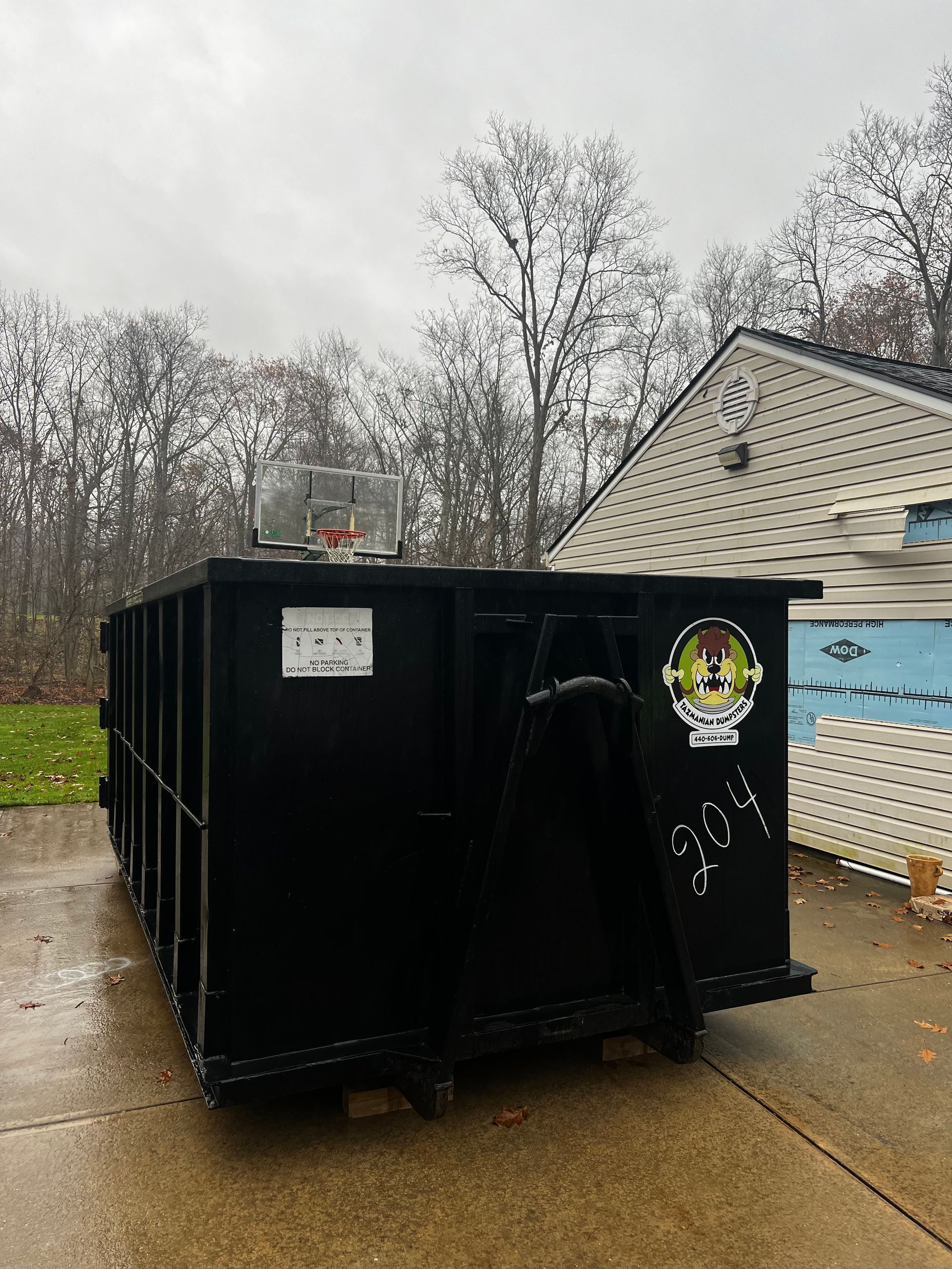 A large black dumpster is parked in a driveway in front of a house.