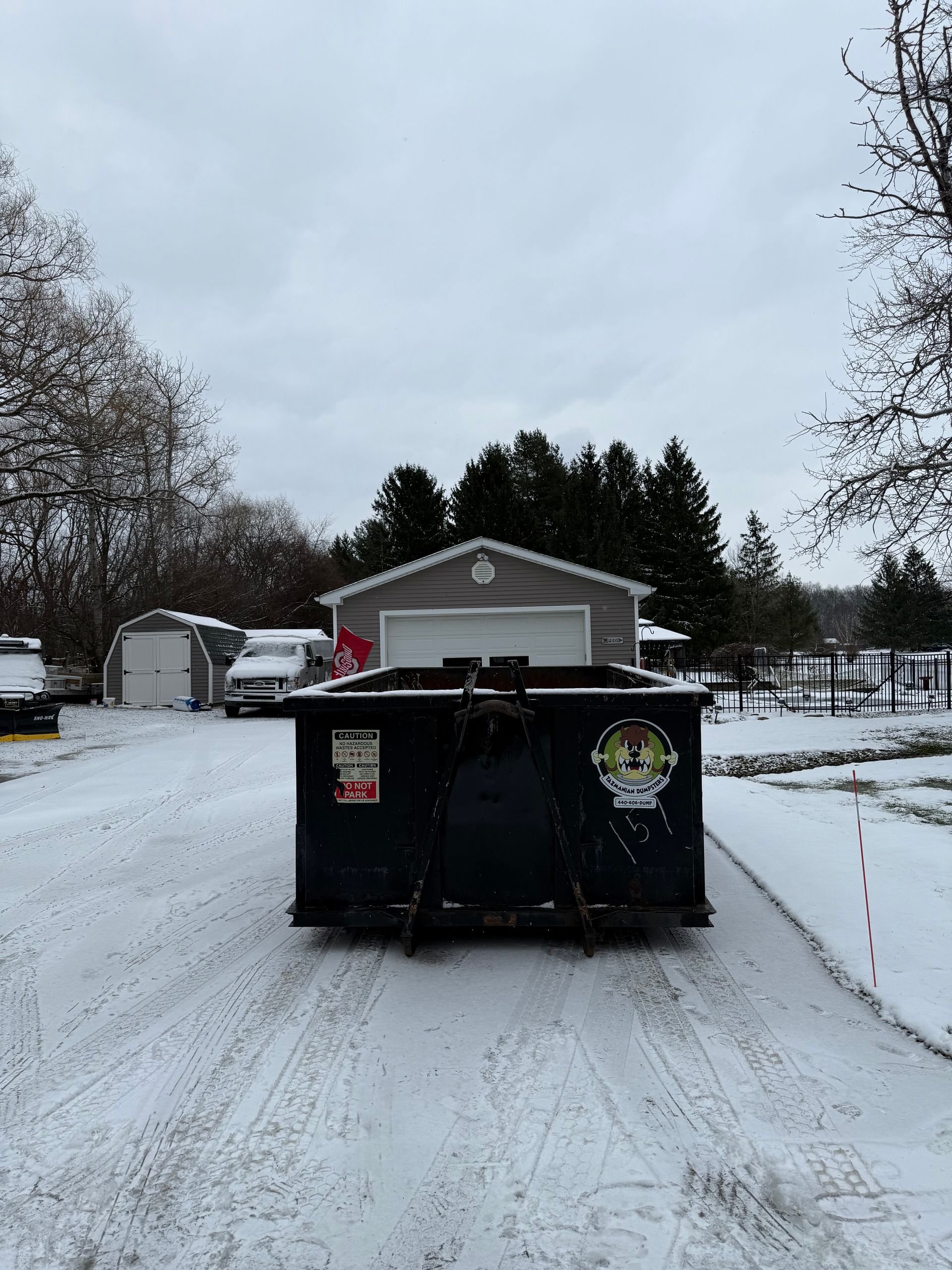 A dumpster is parked in the snow in front of a garage.
