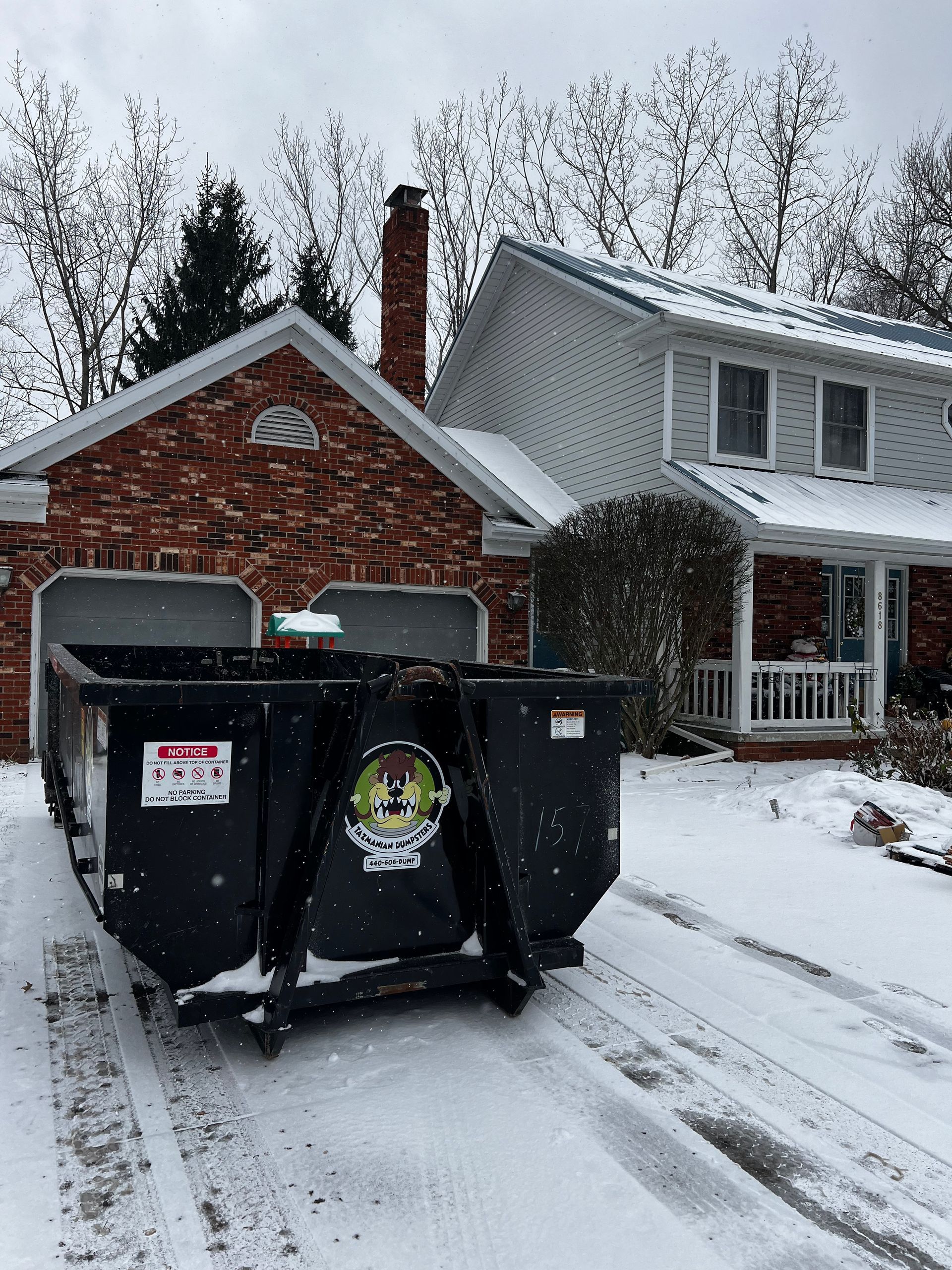 A dumpster is parked in the snow in front of a brick house.