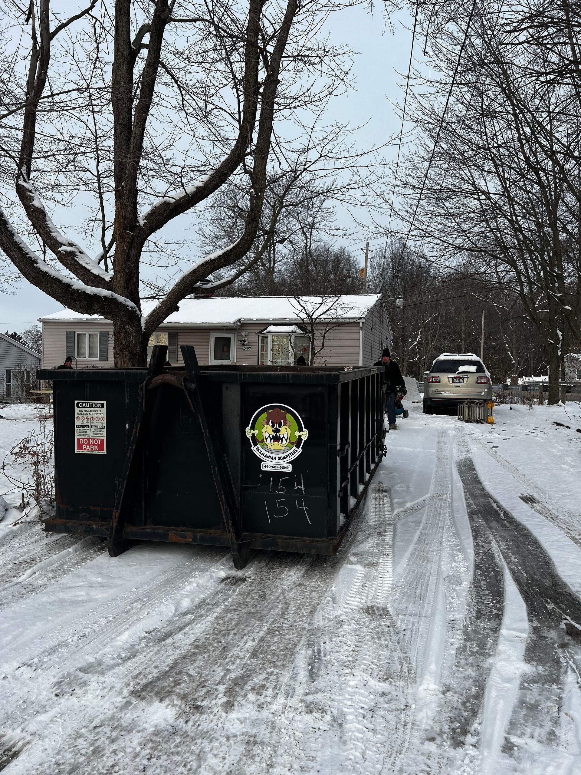 A dumpster is sitting on the side of a snowy road in front of a house.