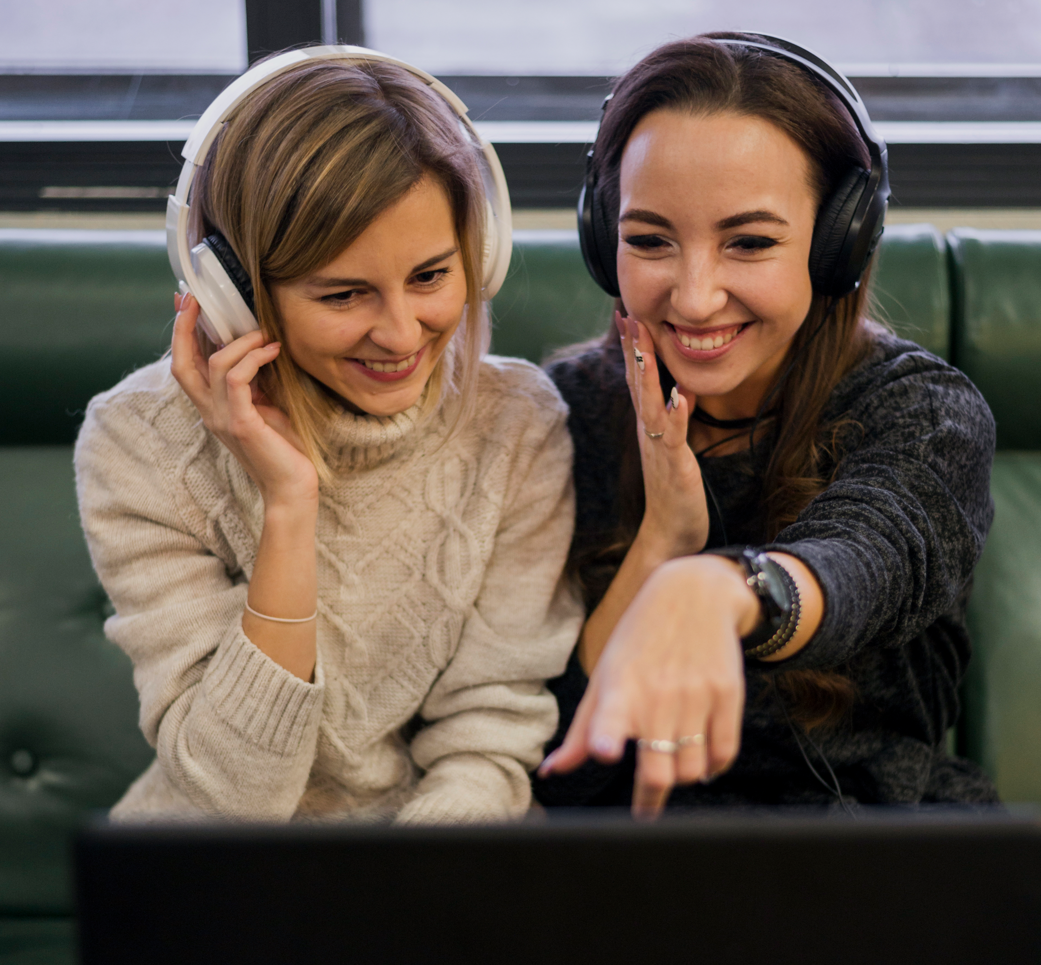 Dos mujeres con auriculares, sonriendo, señalando la pantalla de una computadora portátil. Están en un espacio interior, posiblemente en un salón.