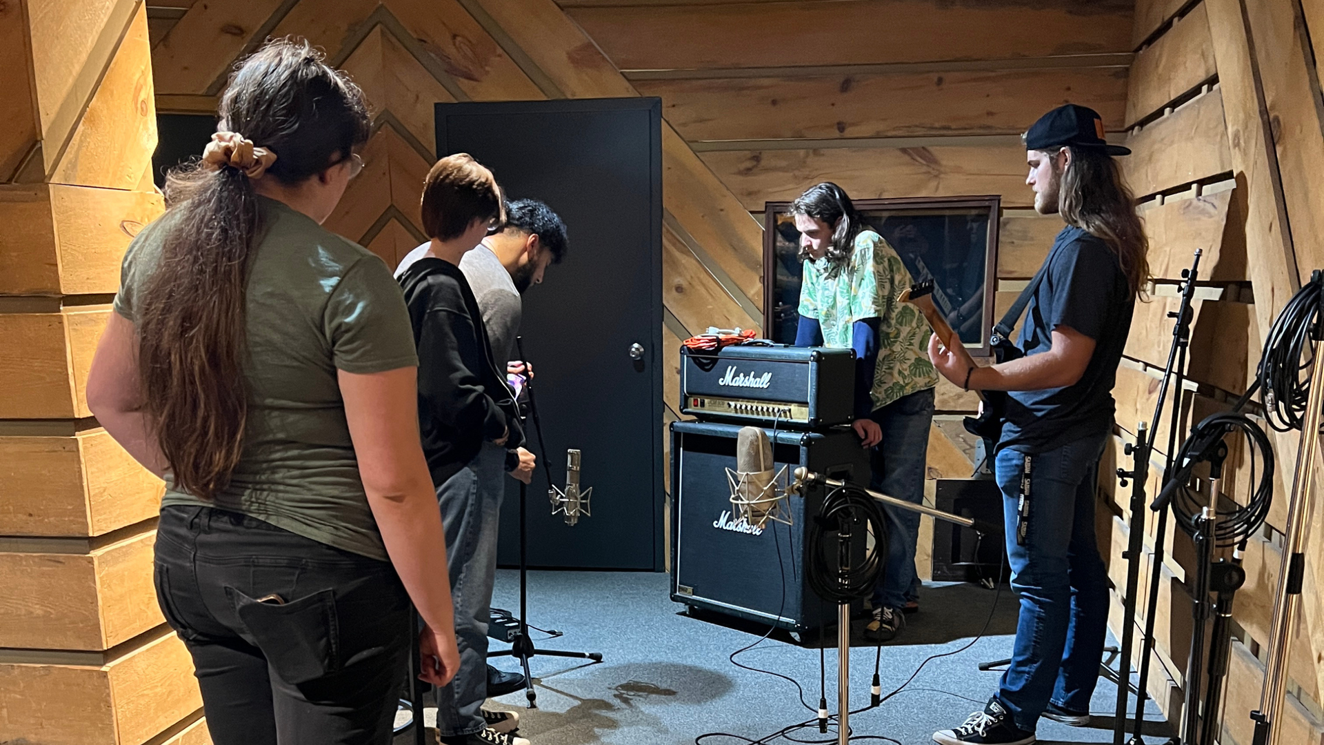 Students in a wood-paneled recording studio. Musicians looking at an amp, some with mics.