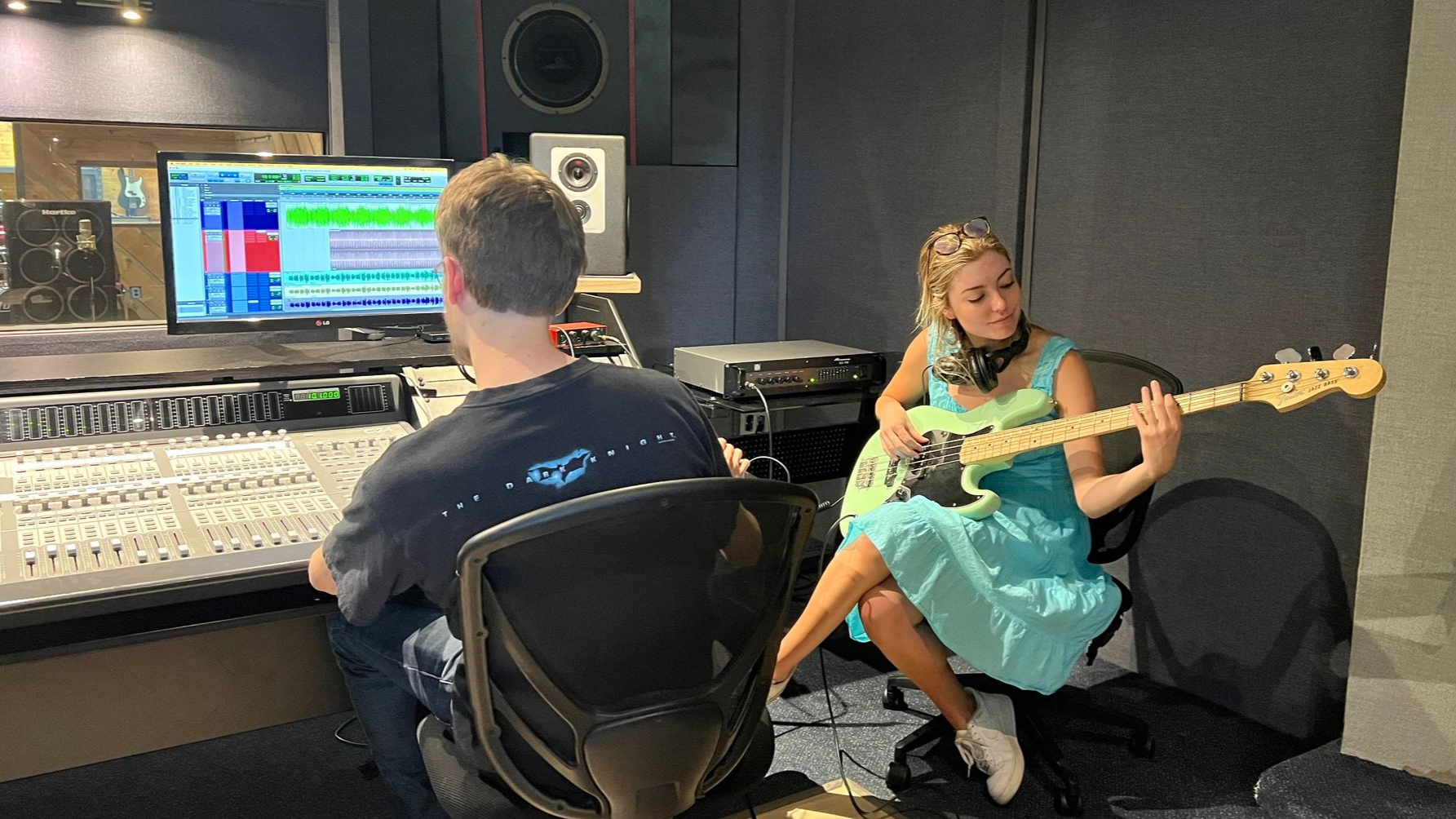 Man at mixing board, woman in blue dress plays bass guitar in recording studio.