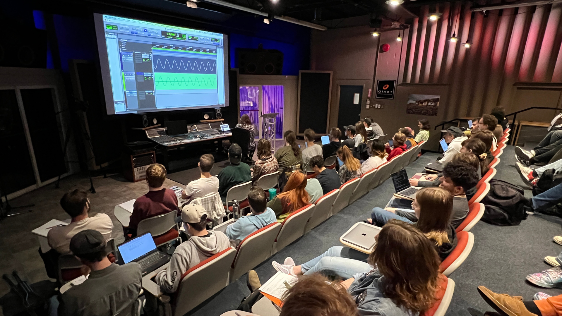 Audience in a music production classroom watches a screen displaying audio software.