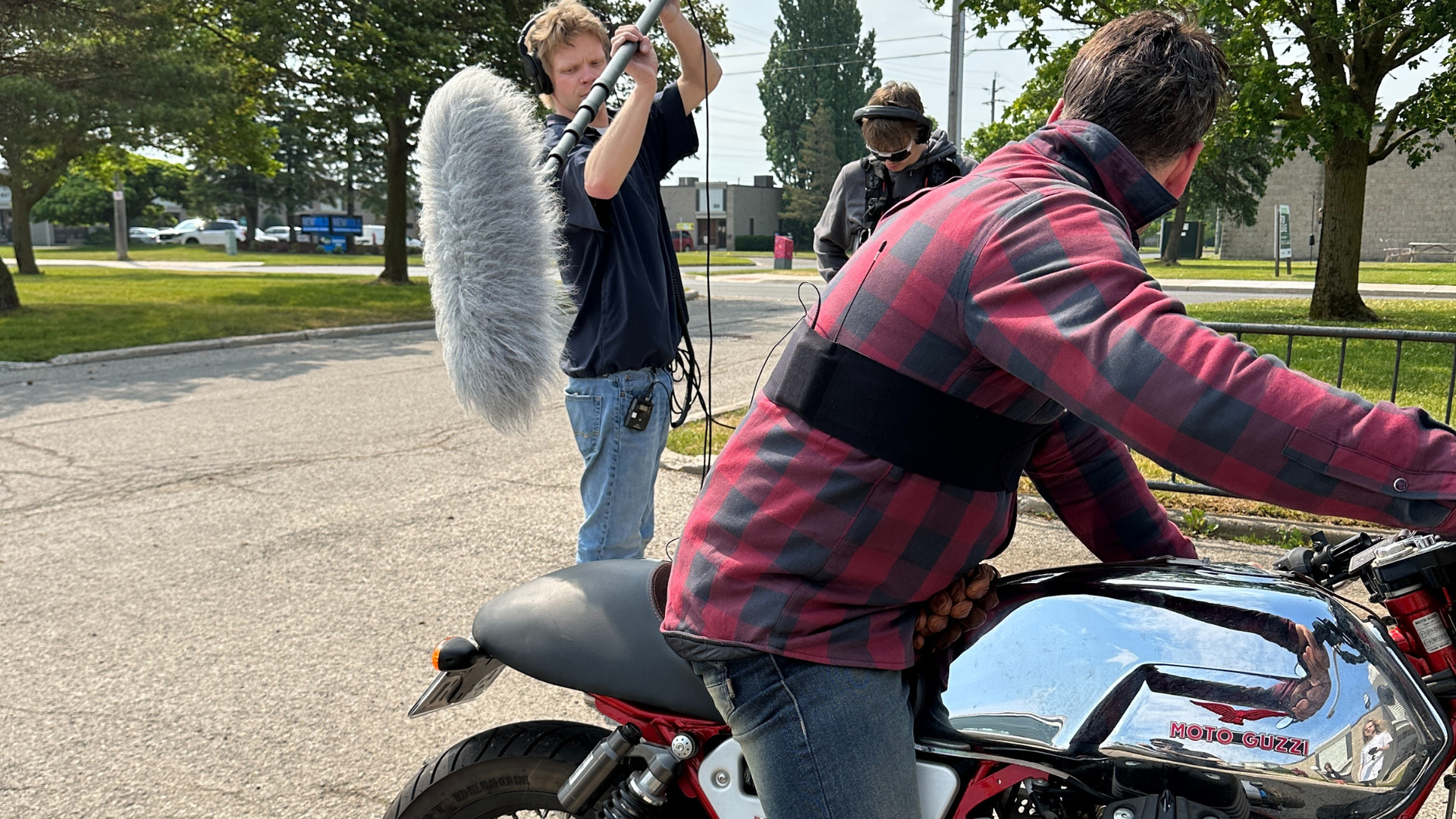 A person on a motorcycle is filmed, with a boom mic and two crew members on a sunny day.