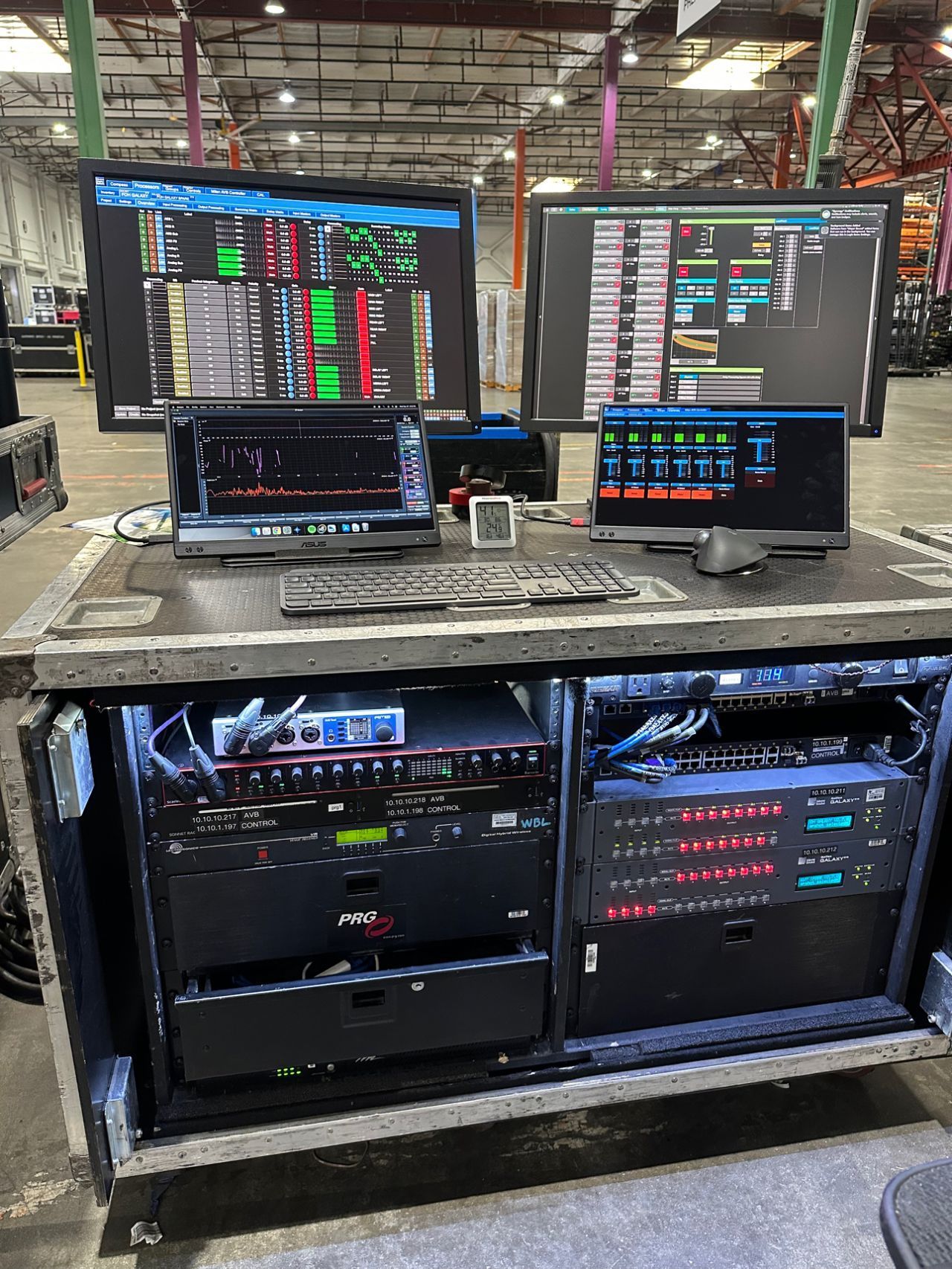 A computer desk with two monitors and a laptop on it in a warehouse.
