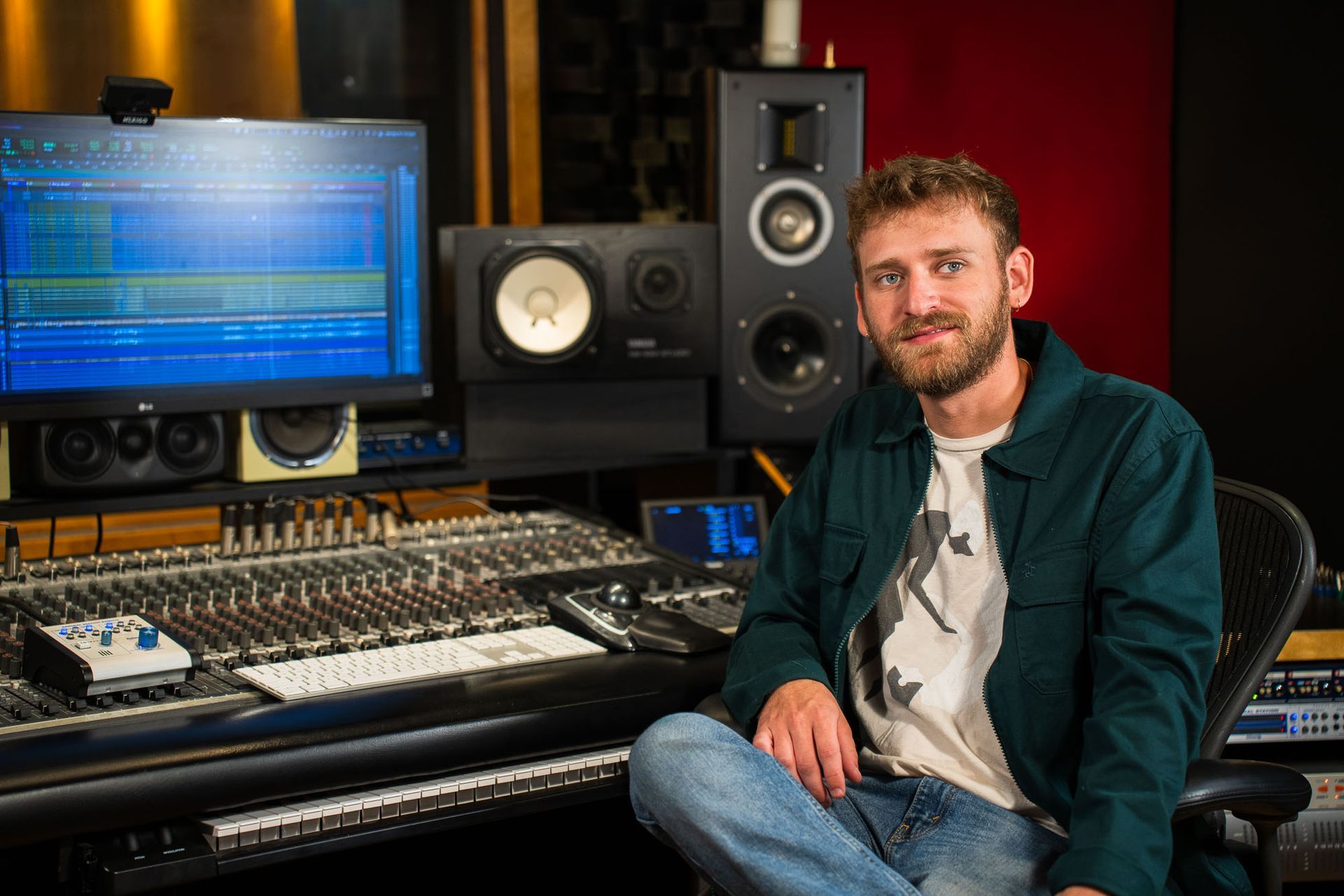 Man in recording studio sits near mixing console, smiling at camera.