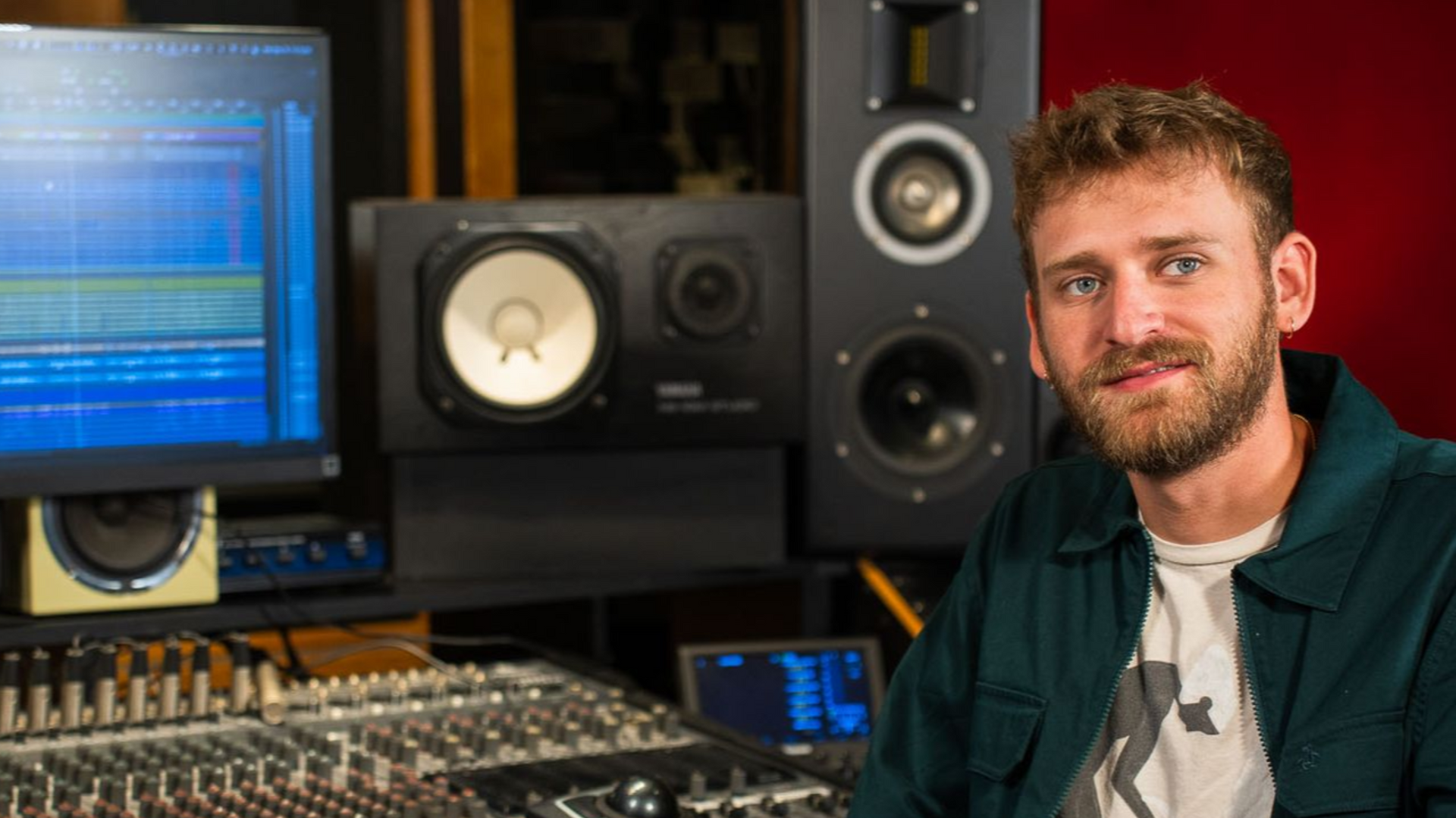 A person with a beard sits at a music studio mixing console, with computer monitors and studio speakers in the background.
