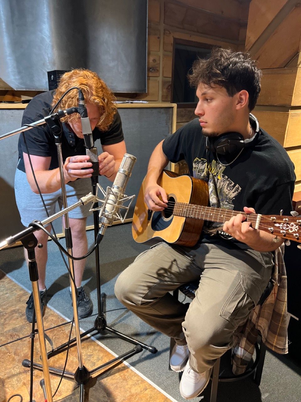 Two men are playing guitars in front of microphones in a recording studio.