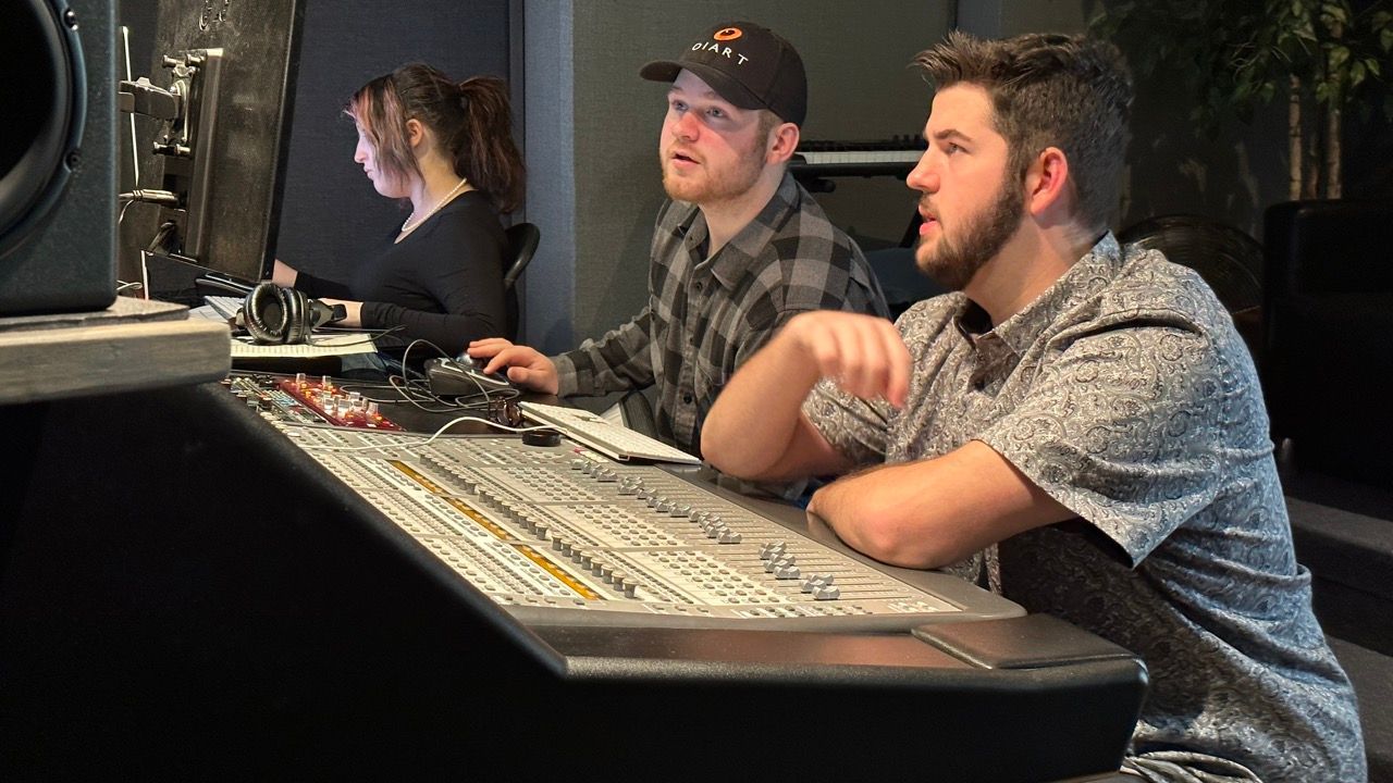 Two men are sitting at a control panel in a recording studio.