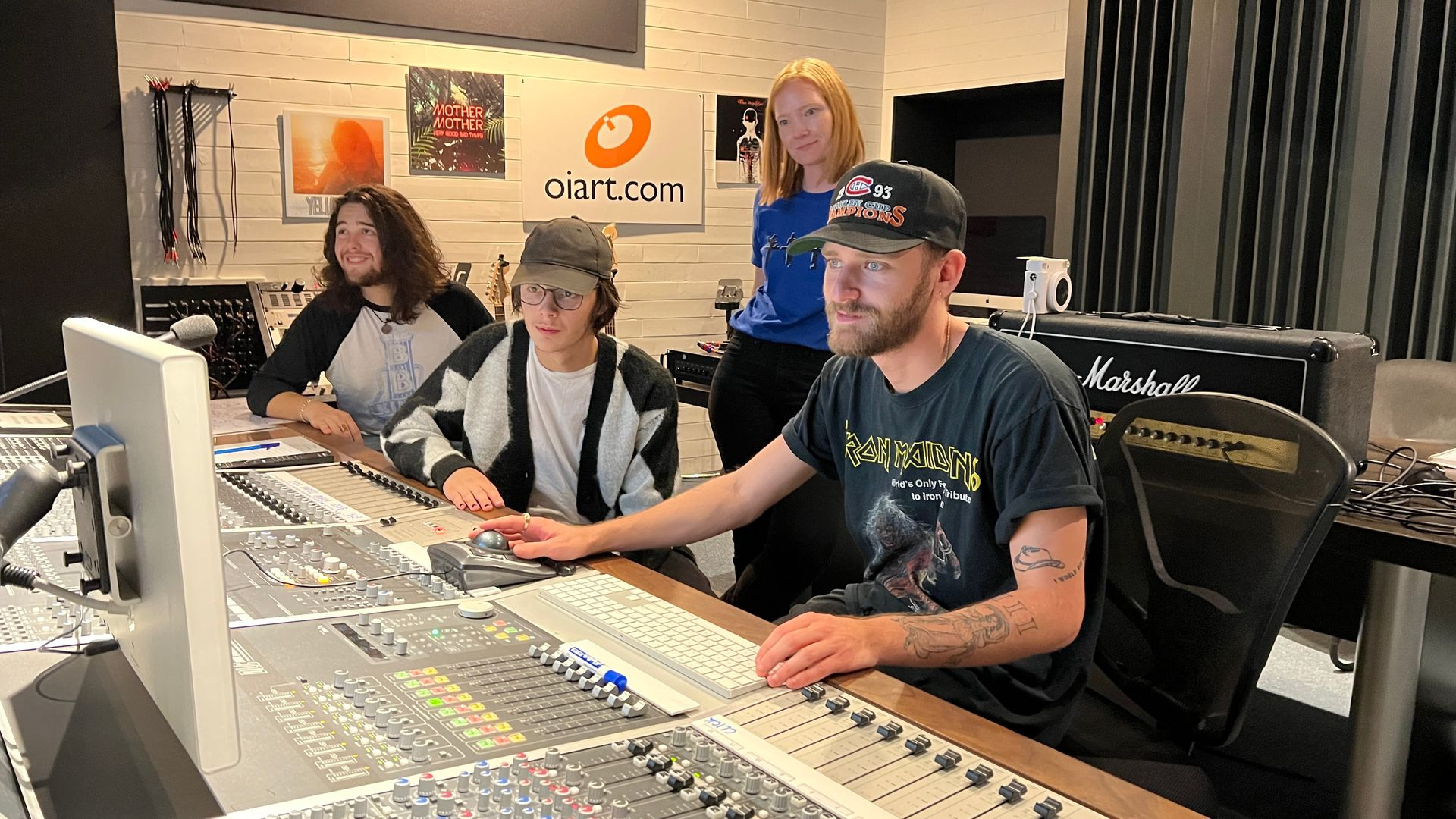 Four people in a recording studio around a mixing console. One man operates the controls while others observe.